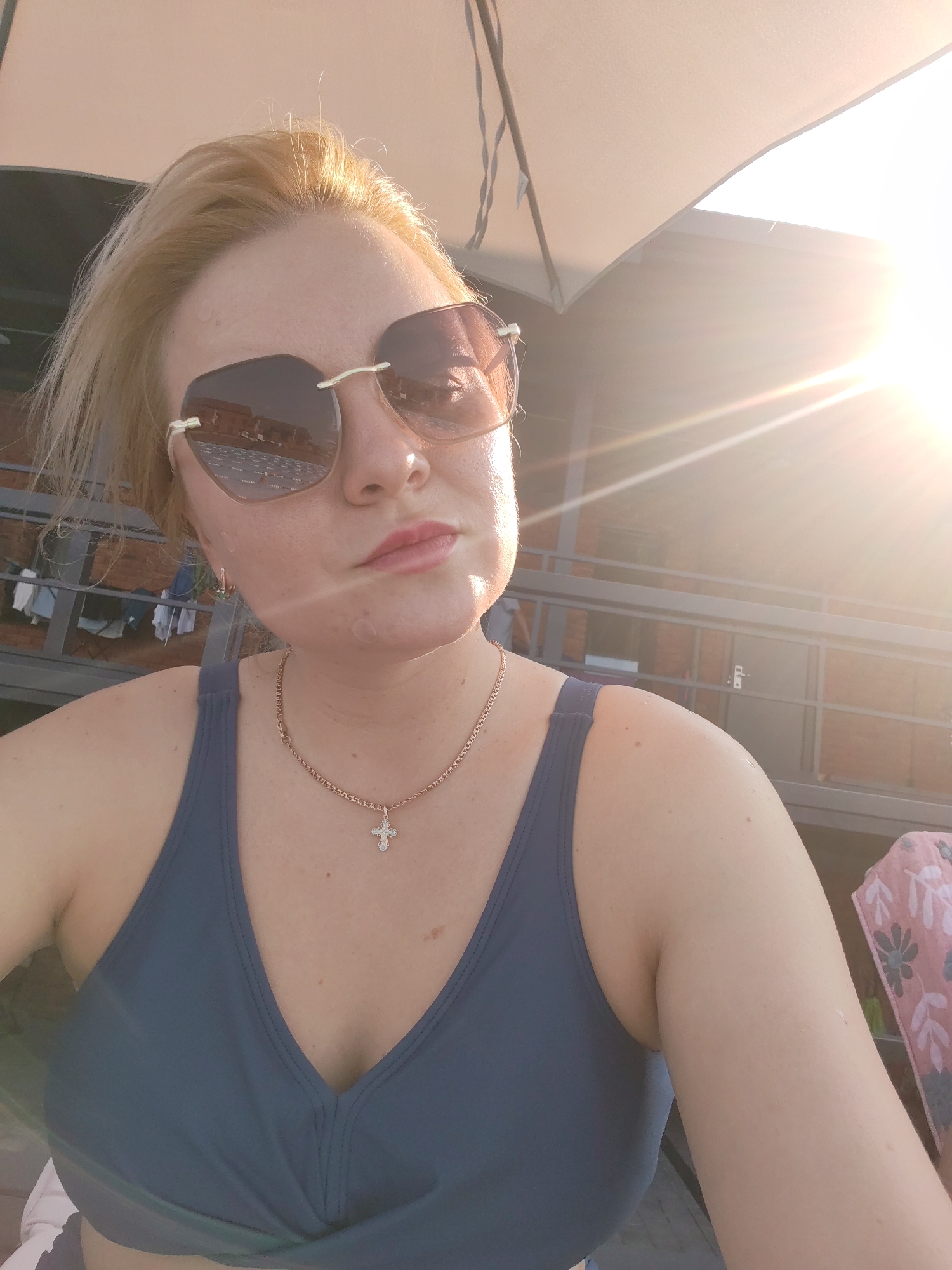 A woman with blonde hair and sunglasses is taking a selfie outdoors. She is wearing a navy blue swimsuit top and a necklace with a cross pendant. The sun is shining brightly, casting a lens flare across the image. In the background, there is a building with a balcony and some laundry hanging on a line. The scene appears to be in Pirogovo, Russia, possibly at a resort or residential area.
