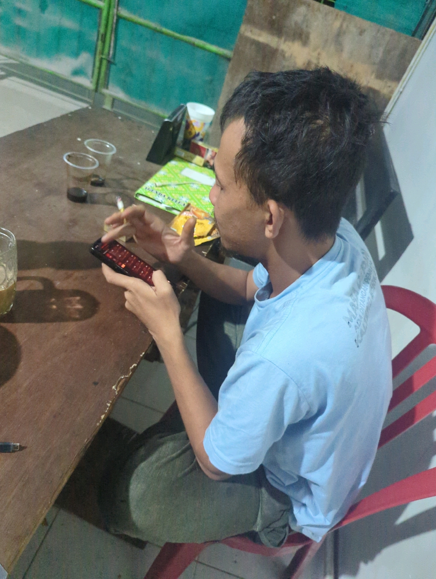 A person is sitting at a brown wooden table, holding a smartphone in their hands and looking at the screen. The person is wearing a light blue t-shirt and dark green pants. On the table, there are two empty plastic cups, a glass with a beverage, a lit cigarette, a package, and a green notebook. The background shows a greenish wall and a wooden partition. The image appears to be taken from a high angle, looking down at the person and the table. The overall scene suggests a casual setting, possibly an outdoor area or a room with limited natural light. The location is identified as Batu Belaman, Indonesia, implying a specific geographical context for this scene.