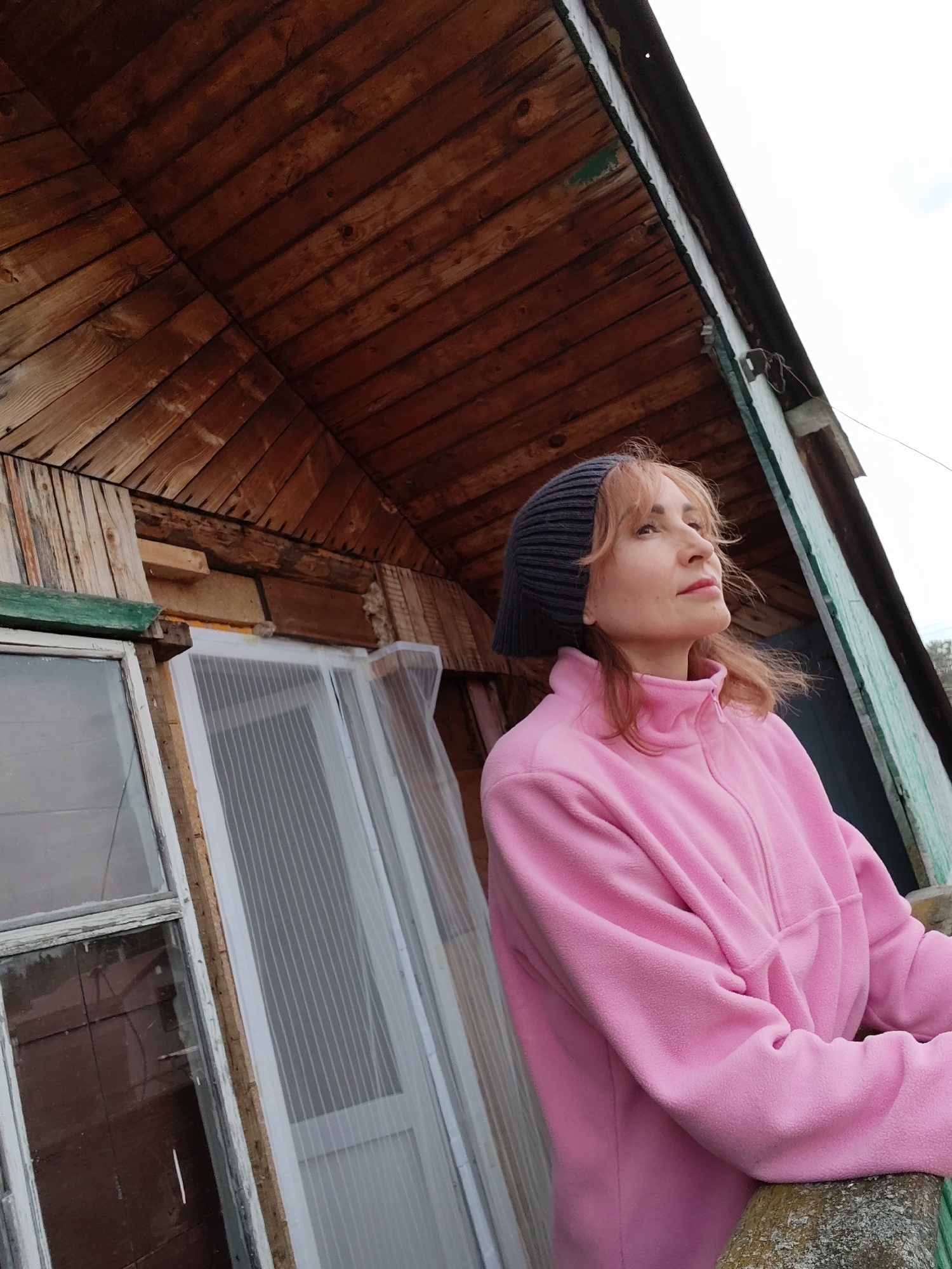 A medium shot shows a woman in a pink fleece jacket and a gray beanie, gazing upwards to her right. She is standing on a weathered, presumably wooden, railing or ledge. Behind her is a rustic wooden building. The wood is stained a deep brown, and the construction appears somewhat haphazard and unfinished. A window frame with partially broken glass and a second window-like structure covered with white fabric hang behind her. The trim of the building is painted light green. The sky is visible in the upper right, suggesting an overcast day. This scene is set in the city of Chaikovsky, Russia.