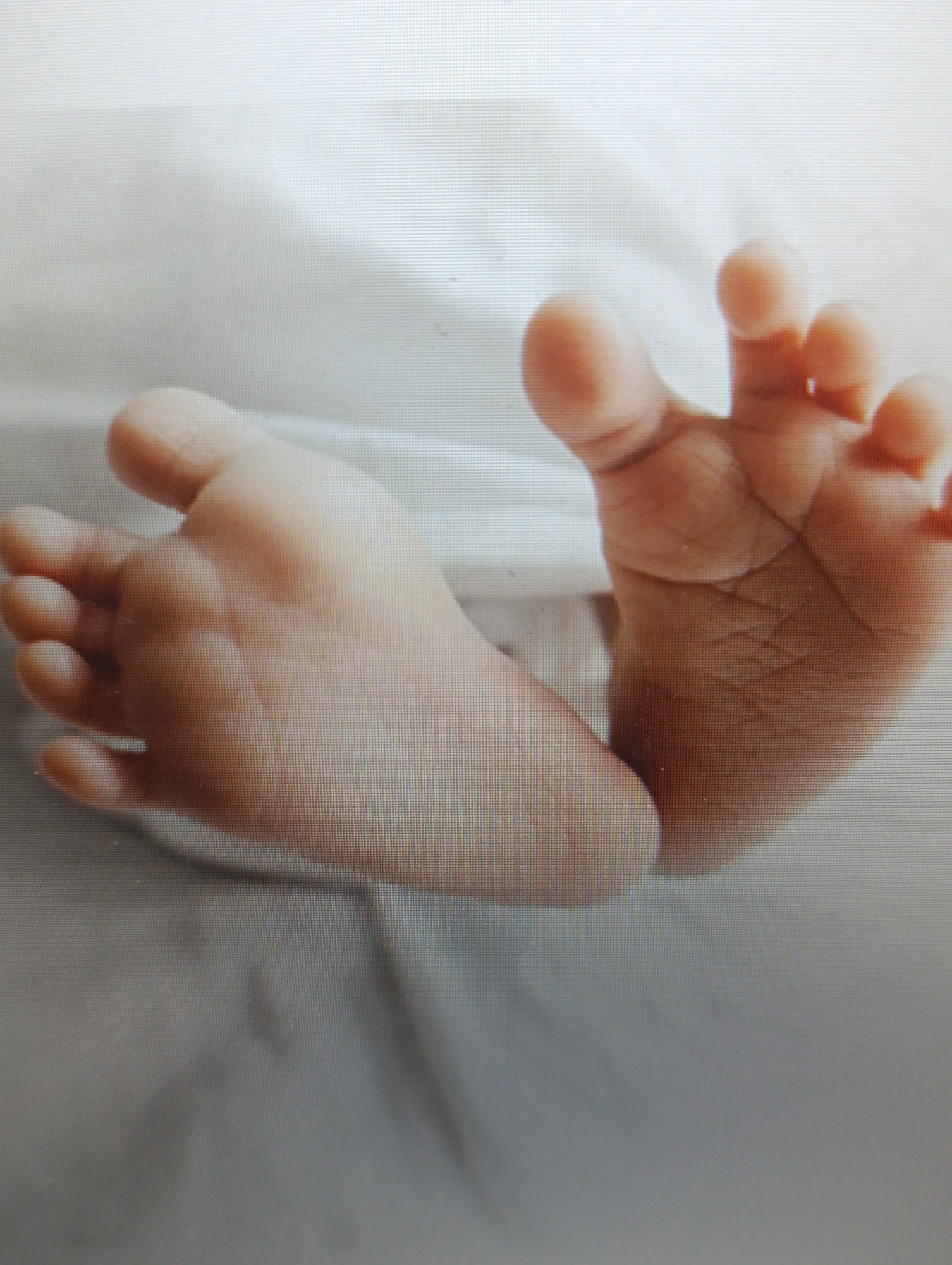 Two baby feet are shown, close-up, on a white, textured surface. The feet are facing upwards and slightly angled away from each other. The left foot is positioned more towards the foreground and left side of the image, while the right foot is further back and to the right. Both feet are light-skinned and appear soft and delicate, with visible toe pads and creases on the soles. The toes of the right foot are slightly spread open, as if reaching or grasping. The lighting is soft and diffused, creating gentle shadows. The background is a plain, white, textured fabric, possibly a blanket or sheet.