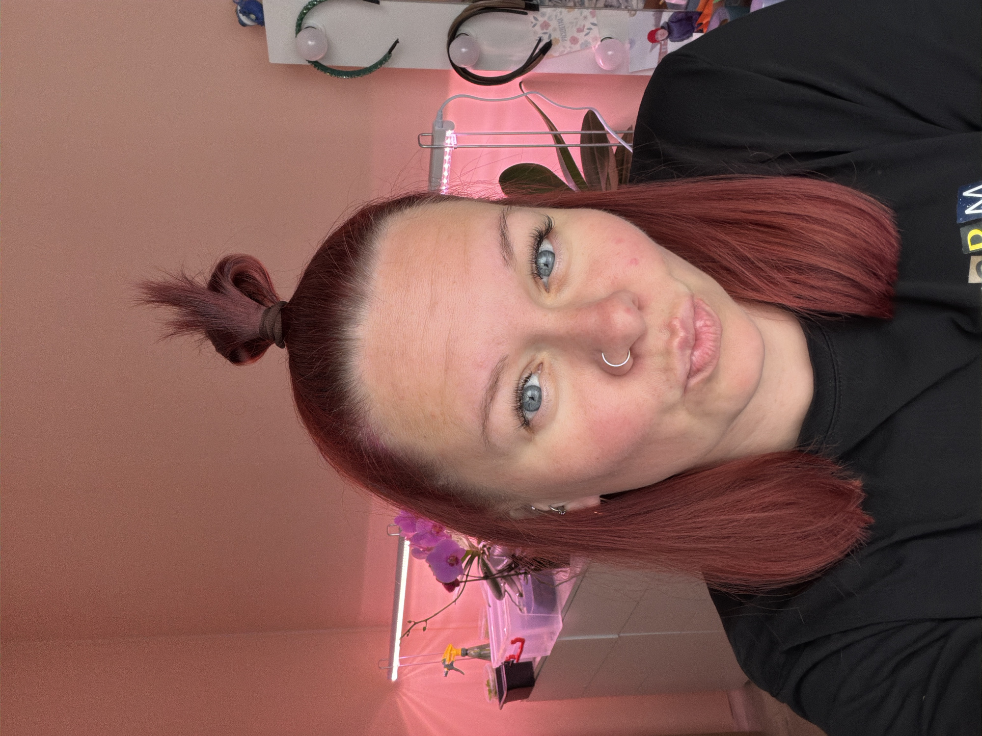 A selfie of a fair-skinned person with red-auburn hair pulled back in a small topknot, wearing a black long-sleeved shirt, and a silver nose ring. The person has blue eyes and is looking directly at the camera with a slight pout. The background is a light pink wall. On the wall, several headbands are attached to a rack, with a plant under artificial light. An orchid plant is also visible below. The location is not explicitly stated in the image. It is impossible to know if the picture was taken in the city of Chaikovsky, Russia.