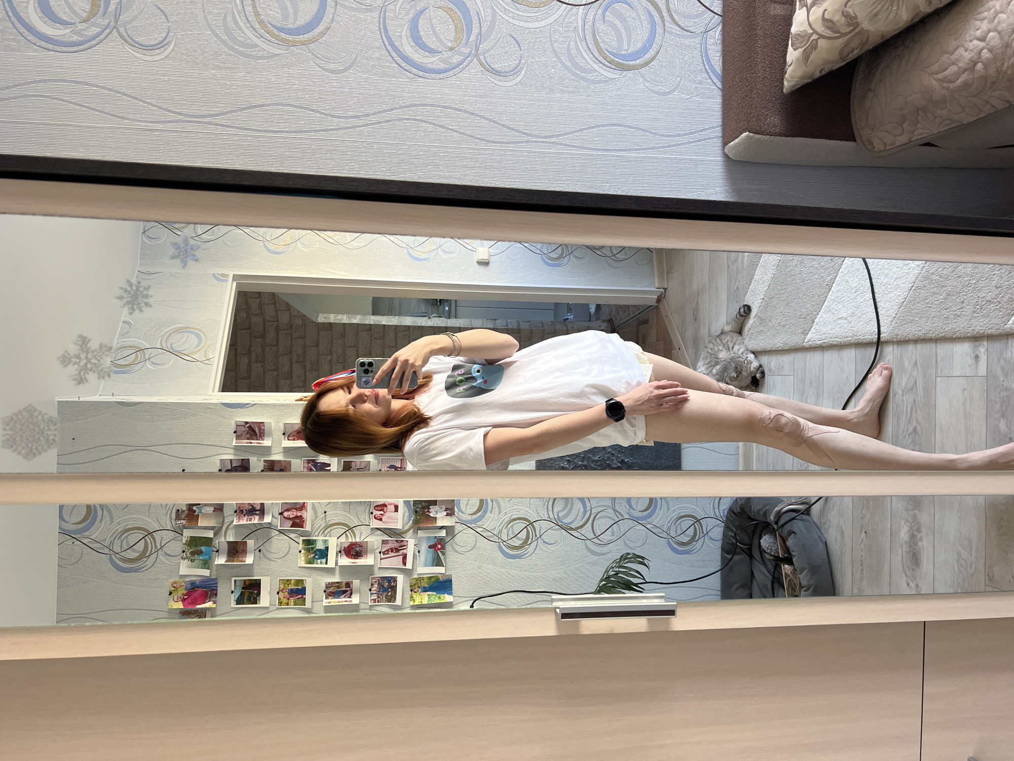 A reflection in a mirror shows a slender, light-skinned woman with auburn hair lying on her side, holding a phone to take a picture of herself. She is wearing a white t-shirt and light-colored shorts. On her left wrist is a black smartwatch. Her legs are extended towards the right, and there are distinct markings or possibly tattoos on her right thigh. In the background, reflected in the mirror, are interior scenes: a wall decorated with photos, a hallway leading to another room, and a pet bed with a grey cat on a grey rug. The mirror itself is framed in black and wood. The location is in Chaikovsky, Russia.