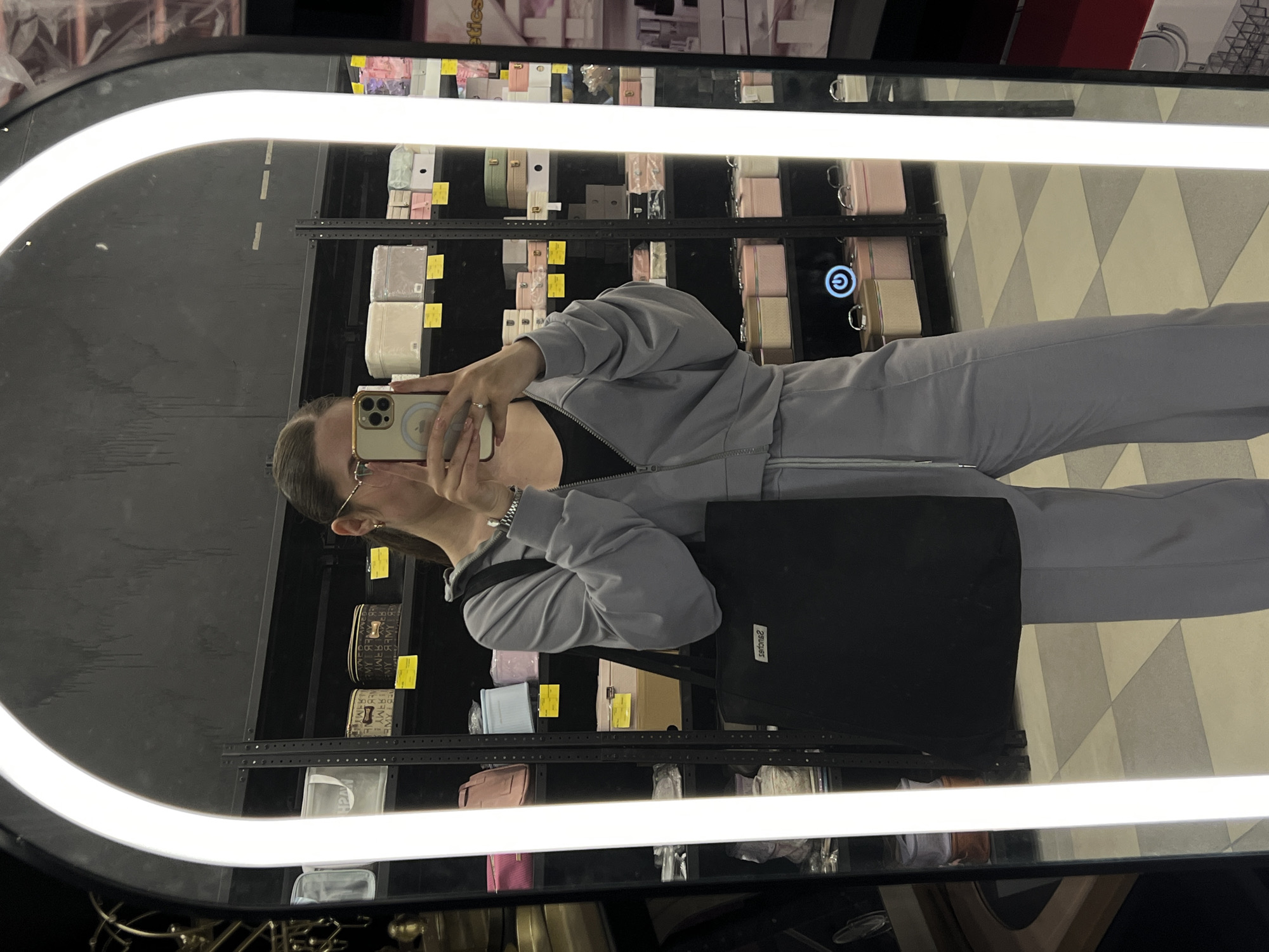The image displays a full-body mirror selfie taken by a person in an indoor commercial setting, likely a store, located in Ternopil, Ukraine. The individual is reflected in a large, upright mirror with an arched top, illuminated by a bright, continuous white LED light strip along its inner edge.
The subject is holding a light-colored smartphone to their face, mostly obscuring their features, though brown hair tied back and clear-framed glasses are partially visible. They are dressed in a light grey or lavender-toned tracksuit, comprising a zip-up jacket worn over a black top, and matching pants. A black tote bag, featuring white, reversed text that appears to read "səuɟəuəs" (likely "sanford" or a similar brand), hangs from their left shoulder. A ring is visible on their left hand.
Behind the person, visible in the reflection, are several retail shelves stocked with numerous packaged items. These items, predominantly in light pastel colors like pink, cream, light green, and white, appear to be small boxes, cases, or pouches, possibly cosmetics, stationery, or fashion accessories. Some items have yellow labels or price tags. Text visible on some shelf items includes "ARM Y" or "ARMY" repeated on small bags and a partial reflection of "etics" (likely from "cosmetics" or similar). A power symbol, a 'G' within an incomplete circle, is reflected on a surface on one of the shelves. The floor visible in the reflection features a light-colored geometric pattern composed of diamond shapes. The scene is brightly lit, indicative of an indoor retail environment.