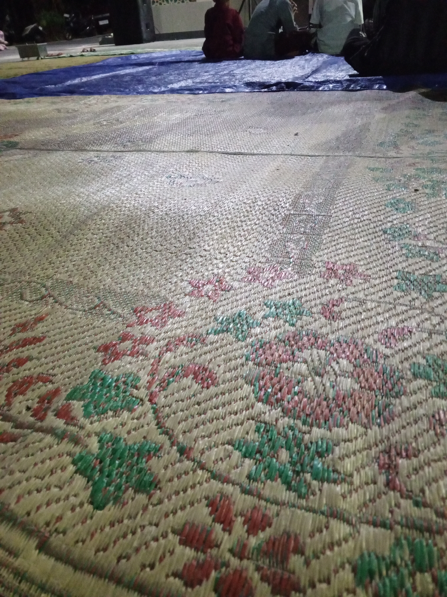 The image shows a close-up view of a woven straw mat with a floral pattern in red and green. In the background, several people are sitting on a blue tarp, which is laid out on a gravelly surface. The setting appears to be outdoors, possibly a communal gathering or event. The overall lighting suggests it might be evening or an overcast day. The location is identified as Lemahbang, Indonesia.