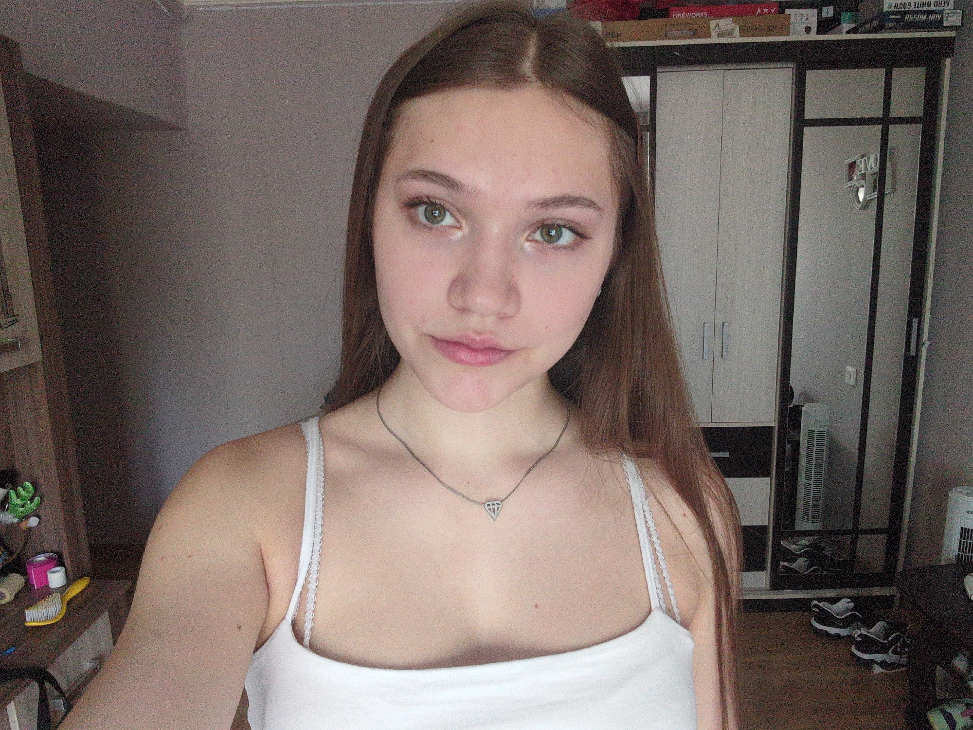 A young woman with long brown hair and green eyes stands indoors, wearing a white tank top and a silver necklace with a diamond pendant. She is positioned in front of a gray wall and a white wardrobe with sliding doors. The room also contains a wooden shelving unit on the left and a mirror with a black frame on the right, reflecting shoes and a fan. Boxes with text such as "FIREWORKS" and "AERO WHITE GUM" are visible on top of the wardrobe. The location is an unidentified interior setting.