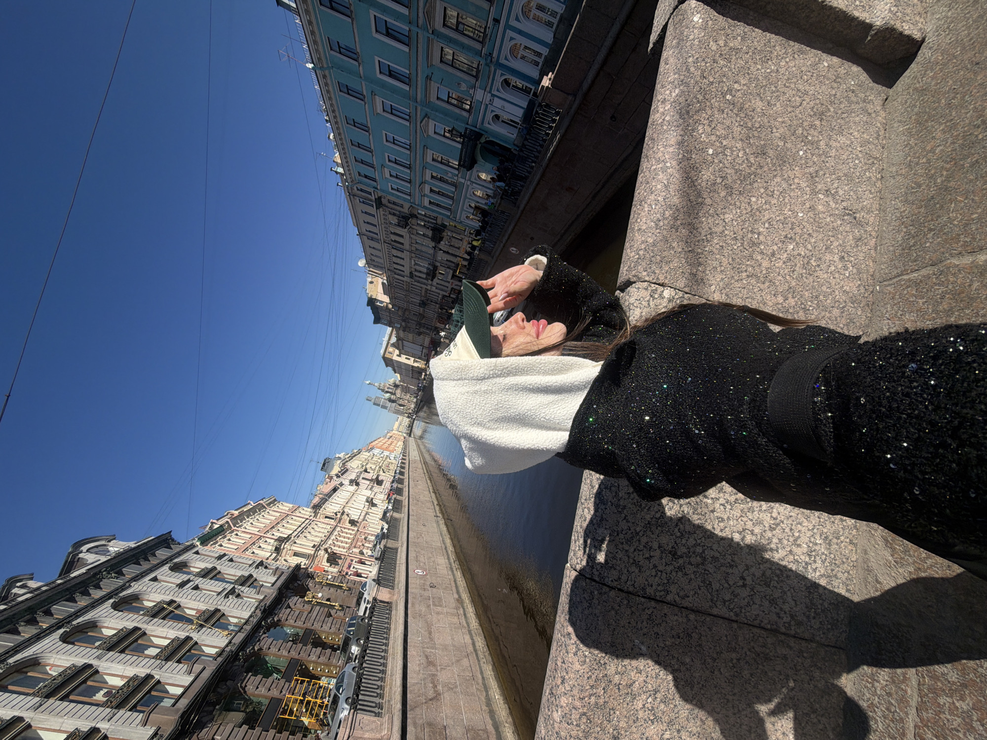 A person is captured in a selfie on a bright, sunny day in Saint Petersburg, Russia, overlooking a canal lined with historic architecture. The image shows the upper torso of a person, likely female, in the foreground on the right. She wears a black, sequined or glittery top and a white, fuzzy-textured collar or scarf. Her head is tilted, and she wears a dark green baseball cap. One hand is raised near her face, possibly shielding her eyes from the sun or adjusting her cap. Her lips are slightly parted.
She appears to be leaning against or standing on a granite bridge balustrade or embankment, which occupies the lower right of the frame. Beyond this, a dark-watered canal stretches into the distance, with an embankment and parked cars visible on the far bank. Rows of multi-story buildings line both sides of the canal. On the left side, buildings display varied architectural styles, some light-colored and others with intricate details. Across the canal, on the right bank, a distinctive series of blue-hued buildings with white trim and numerous windows are prominent. In the far distance, down the canal on the left, the domes or spires of a cathedral are faintly visible.
The sky is a clear, vibrant blue, indicating excellent weather. Thin power lines crisscross the sky. The bright sun casts sharp, dark shadows of the person and the granite balustrade on the ground. No discernible text is visible on buildings or the person's attire, though faint, illegible white lettering is present on the cap.