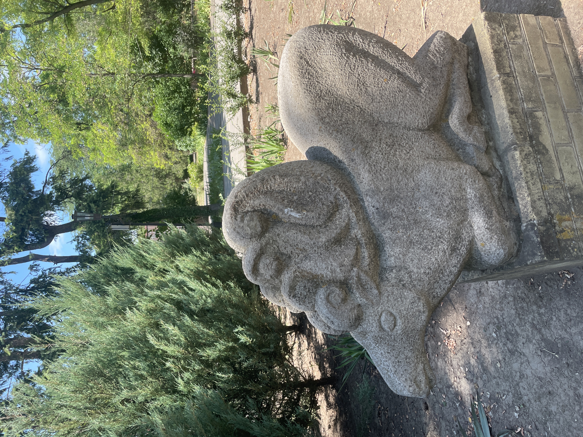 A stone sculpture of a stylized ram or sheep is situated in a park-like setting. The sculpture is predominantly grey and appears to be carved from rough-textured stone, showing some weathering and discoloration. It is positioned on a base made of brickwork.

Behind the sculpture, a dense green juniper bush dominates the left side of the frame, with its branches reaching upwards and outwards. Further back, a path or walkway is visible, lined with smaller green bushes and some dry leaves on the ground. The background consists of a variety of trees with lush green foliage, creating a natural, somewhat wild environment. The sky above is blue with scattered white clouds.

This sculpture is located in the city of Azov, Russia, within the territory of the Azov Museum-Reserve, specifically near the Azov Fortress.