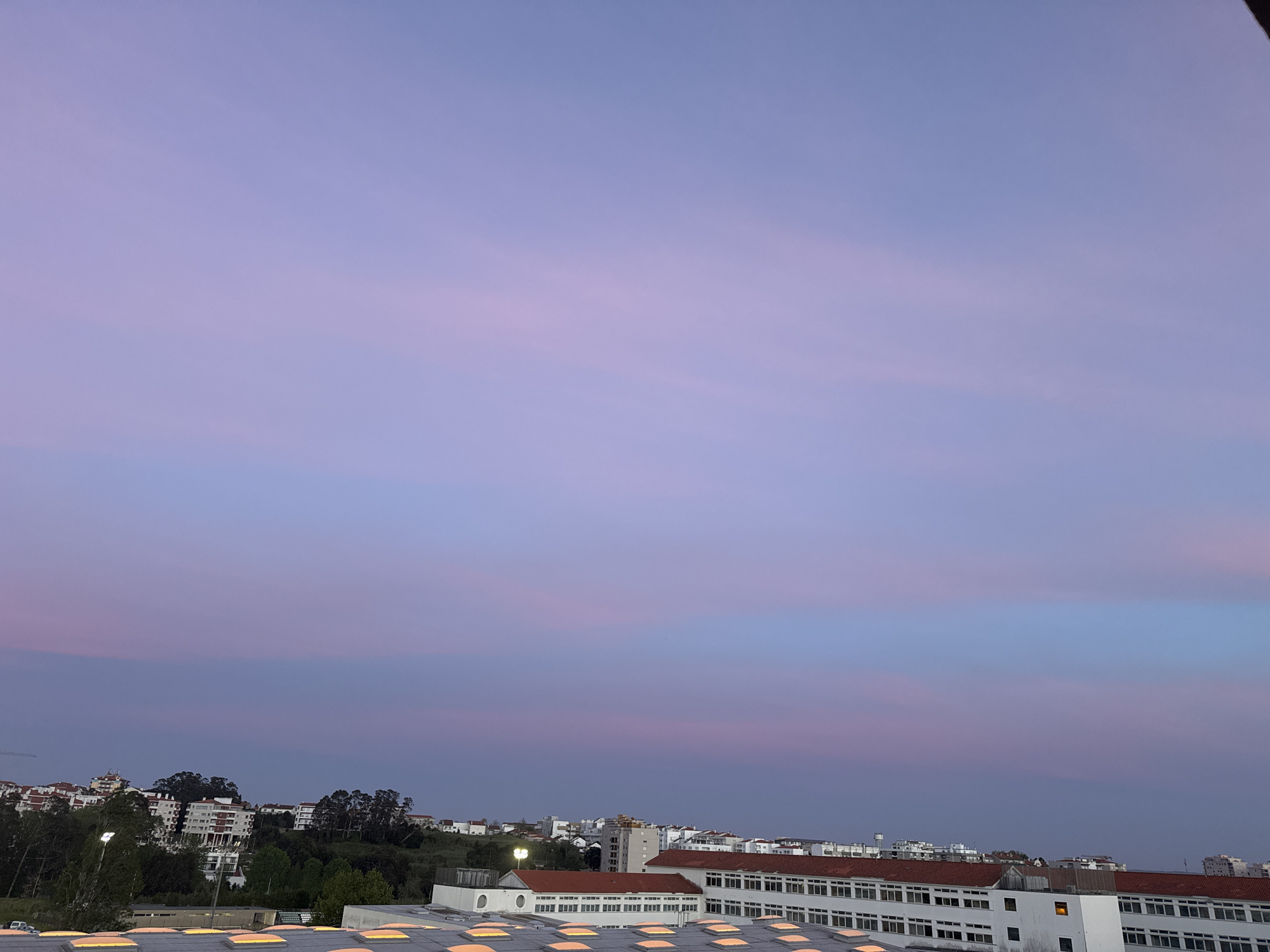 The image captures a serene, expansive view of a town or city at dusk, looking out over a blend of urban structures and natural elements. The most prominent feature is the sky, which fills the upper two-thirds of the frame. It displays a beautiful twilight gradient, transitioning from soft pastel pinks and purples near the horizon to deeper shades of blue-purple higher up. Thin, wispy clouds are visible across the sky, catching and reflecting these ethereal hues.
In the mid-ground and foreground, a sprawling landscape of buildings and trees is visible. On the left, residential buildings with light-colored facades and terracotta roofs are nestled among lush green hills and dense trees, suggesting a suburban or less dense area. A bright streetlamp is visible among the trees on the left. On the right, larger, possibly commercial or institutional buildings with white walls and reddish-brown roofs extend across the frame. Some buildings show illuminated windows, hinting at the evening hour. The foreground features the roof of a large building, characterized by numerous square or rectangular skylights arranged in neat rows, reflecting the ambient light of the sky.
No people are visible in the scene. The weather appears calm and clear, with only light cloud cover. The overall impression is one of tranquility and peace as daylight fades, consistent with an evening setting in a place like Figueira da Foz, Portugal, given the architectural style. There is no visible text in the image.