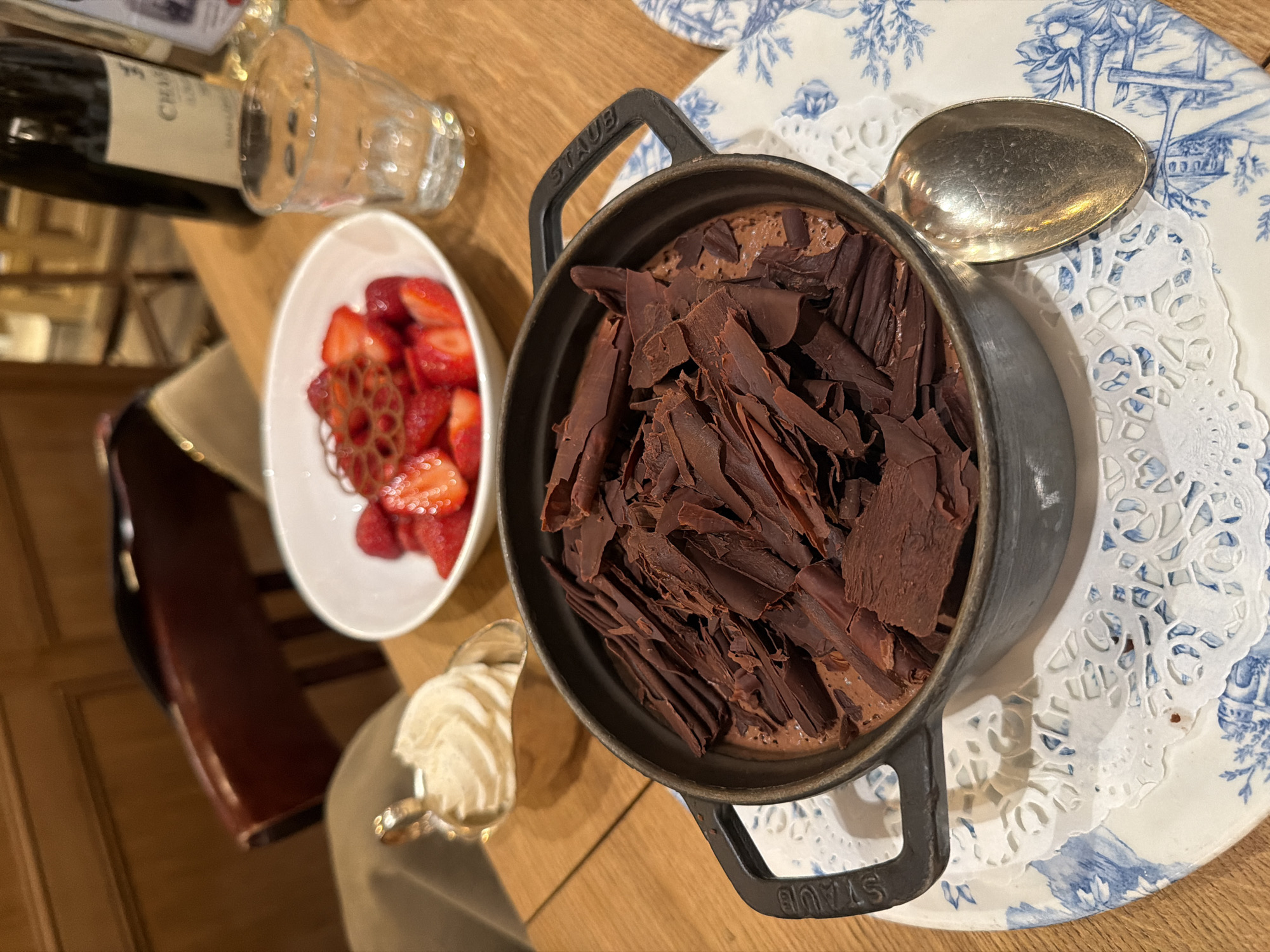 The image depicts a table setting, likely in a restaurant. A small, dark Staub pot filled with chocolate mousse and topped with chocolate shavings sits on a blue and white patterned plate, which is lined with a white lace doily. A silver spoon rests on the plate beside the pot. A small white bowl containing sliced strawberries and a piece of lotus root is positioned to the left of the dessert. A small silver spoon containing whipped cream is also present. A bottle of wine and a glass are situated in the upper left corner of the image. The table is wooden. There is no definitive evidence of Paris, France.