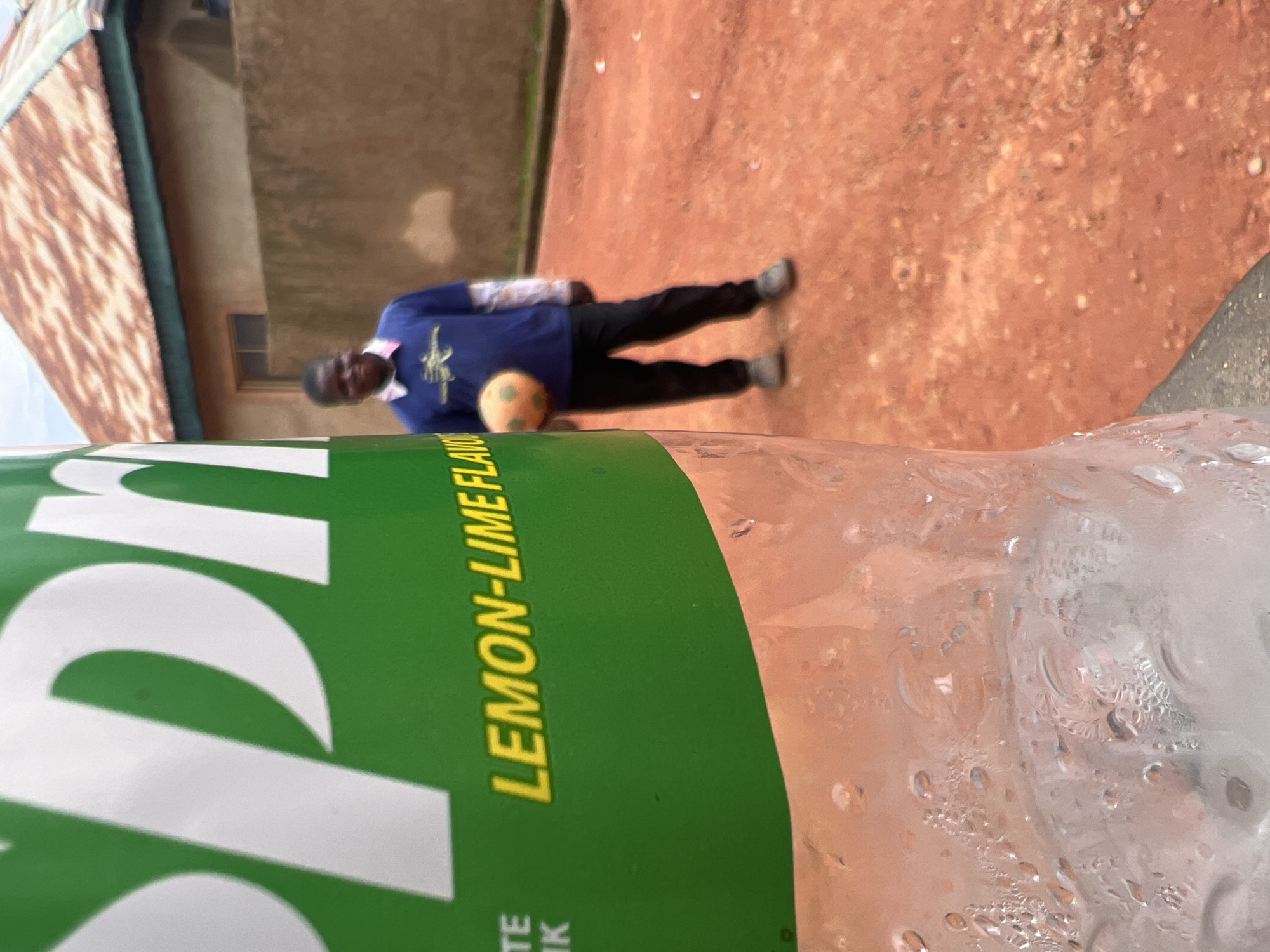 In Jos, Nigeria, a person stands outdoors near a building with a corrugated iron roof. The ground appears to be made of red earth. The person is wearing a blue long-sleeved shirt with a logo and black pants. They are holding a small, round, orange object, possibly a ball. In the foreground, a portion of a plastic bottle is visible, with condensation on its surface. The bottle has a green label with white lettering. Visible text on the label reads "LEMON-LIME FLAVOR" and partially visible text below reads "TE K". The lighting suggests it is daytime, and the overall scene is casual and outdoors.