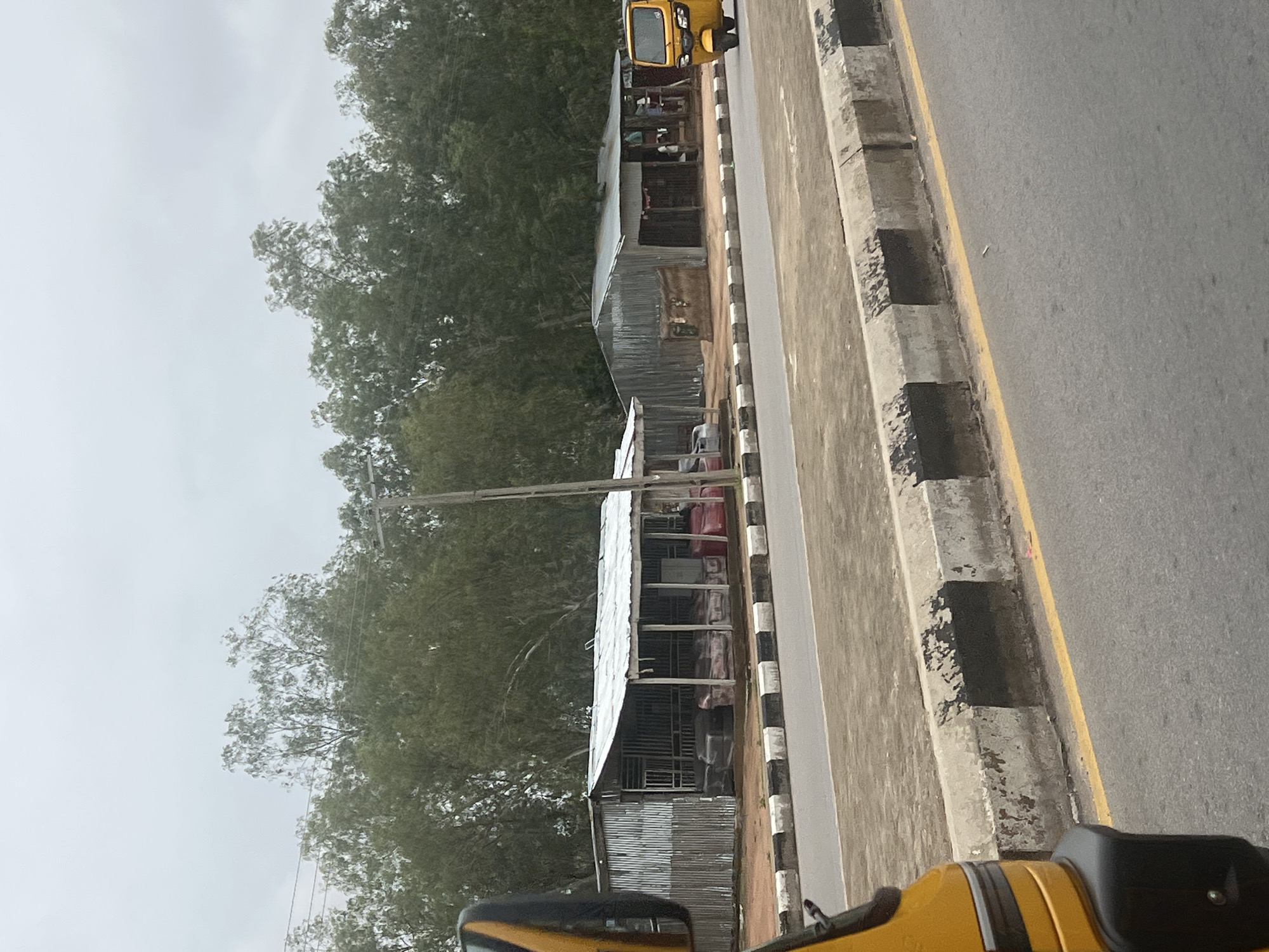 In Jalingo, Nigeria, along a paved road bordered by a concrete curb with black and white stripes, a row of corrugated metal structures with tin roofs serves as shops. In the foreground, the yellow front fender and wheel well of a vehicle are visible on the left. Further down the road, a yellow auto-rickshaw is parked in front of one of the shops. The shops themselves appear to be open-air, with some displaying items for sale, such as what seem to be mattresses or cushions. Behind the shops, a dense line of tall, leafy trees forms a backdrop under an overcast sky. A utility pole is also present between the shops and the trees.