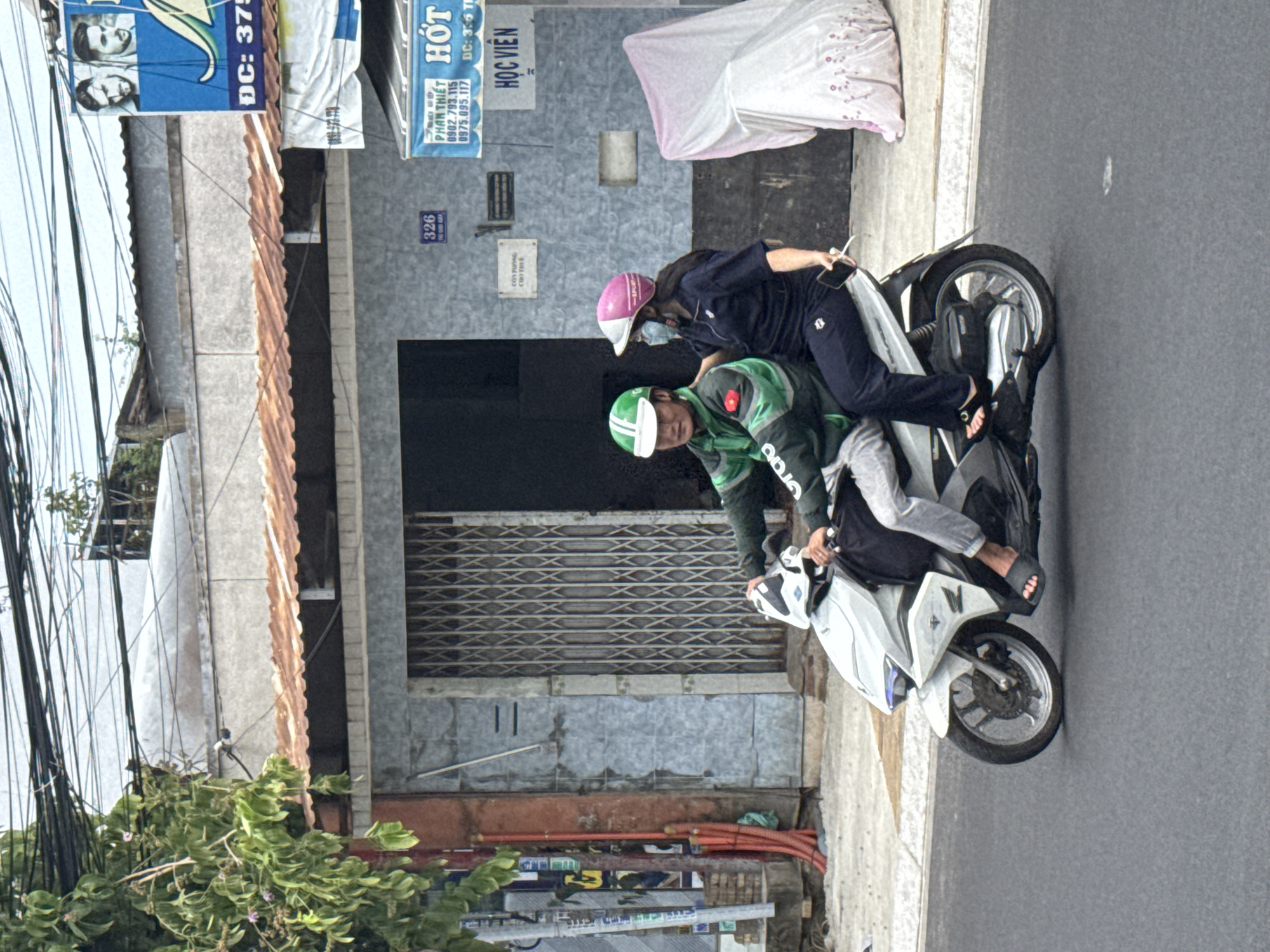 Two individuals are riding a white motorcycle on a paved street in Phú Thủy, Vietnam. The person in the front is driving, wearing a green jacket with a white and green helmet, and a white visor. The passenger in the back is wearing a dark blue outfit and a pink and white helmet, and is holding a smartphone. The motorcycle has a black bag attached to the back. In the background, there is a building with a gray corrugated metal roof, a closed metal gate, and various signs. One sign, partially visible, has Vietnamese text and the word "HỌC VIÊN" (student/academy). Another sign above it reads "PHAN THIET" and displays phone numbers. A third blue sign features images of two men and the text "DC: 375". The environment appears to be an urban or semi-urban setting, with overhead electrical wires visible against a cloudy sky. There are also trees with green foliage to the left of the scene, and a pile of orange pipes on the ground.