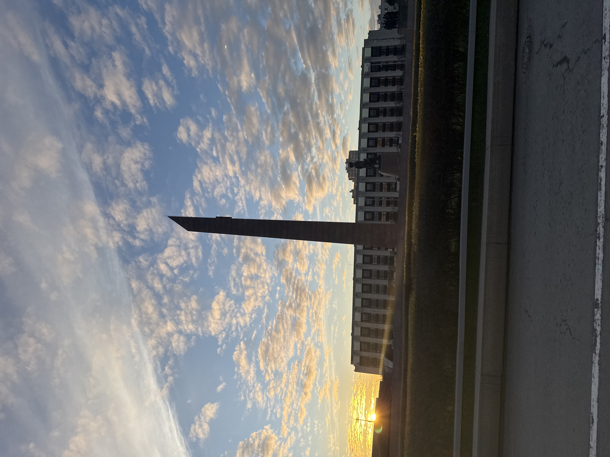 The image depicts a vertical composition with a view of Saint Petersburg, Russia. A tall, dark monument with a sharp, flat top dominates the left side of the frame, reaching into a sky filled with numerous white and yellow clouds against a blue backdrop. To the right of the monument, there's a building with multiple rows of windows. A green hedge runs along the base of the building and monument. A sidewalk and paved road occupy the right side of the image. The sun appears to be setting at the bottom right, casting a warm glow. No people are visible.