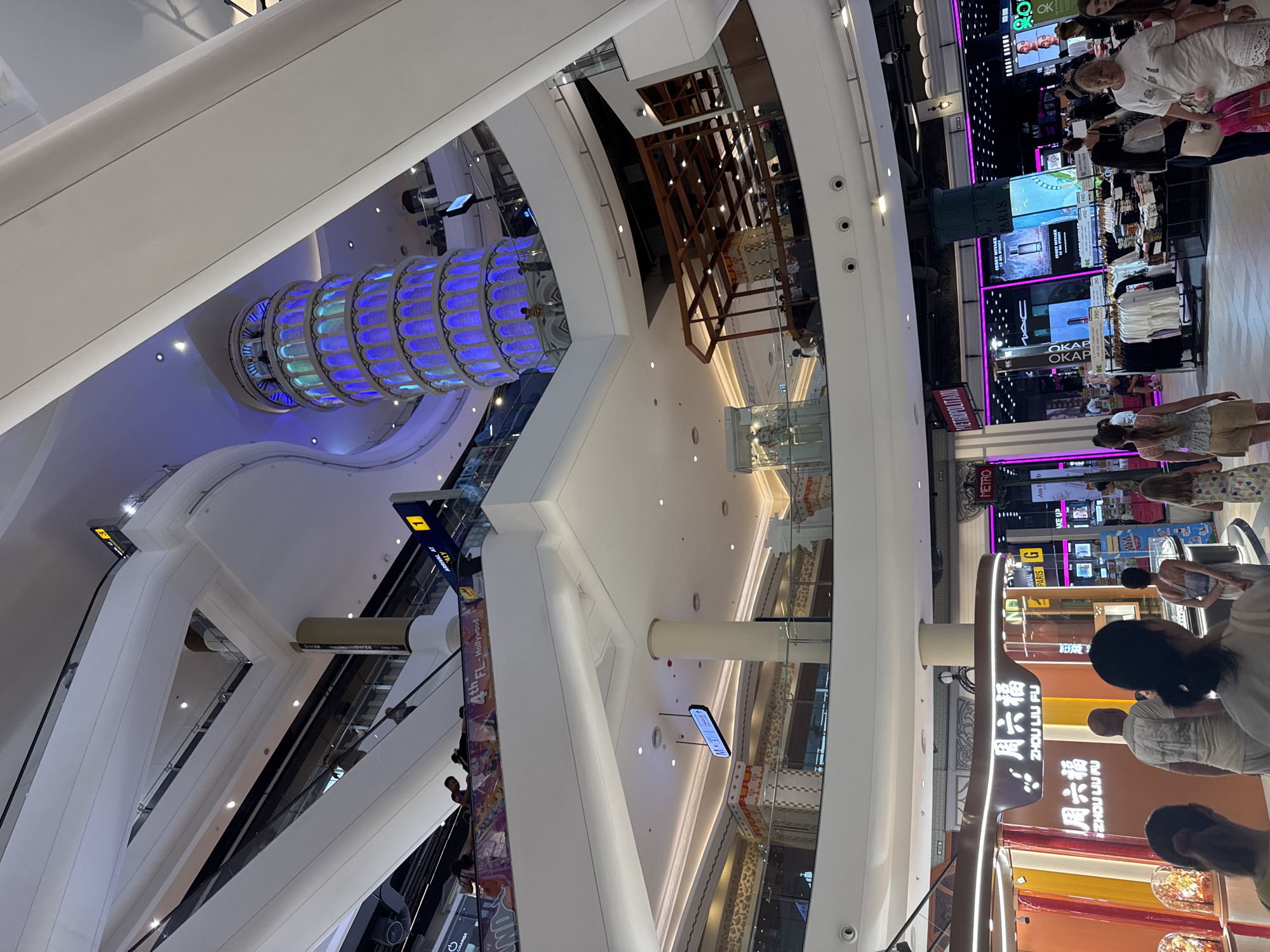 This image shows the interior of a large, multi-level shopping mall in Pattaya City, Thailand. The architecture is modern and airy, with white, curved structures dominating the upper levels. A prominent feature is a replica of the Leaning Tower of Pisa, illuminated with blue and green lights, situated in the center of the mall.
On the lower levels, various shops and escalators are visible. Signs in English and what appear to be Asian languages are displayed, including "ZHOU LIU FU" and "METROPOLITAN." People are seen walking, shopping, and using the escalators. The lighting is a mix of ambient mall lighting and spotlights, creating a bright and vibrant atmosphere.
In the foreground, several individuals are captured from behind, suggesting the photographer is among a group of shoppers. To the left, a jewelry store with display cases containing illuminated objects is visible. To the right, clothing racks with various garments are displayed outside stores. The overall scene depicts a busy commercial environment within a contemporary shopping complex.