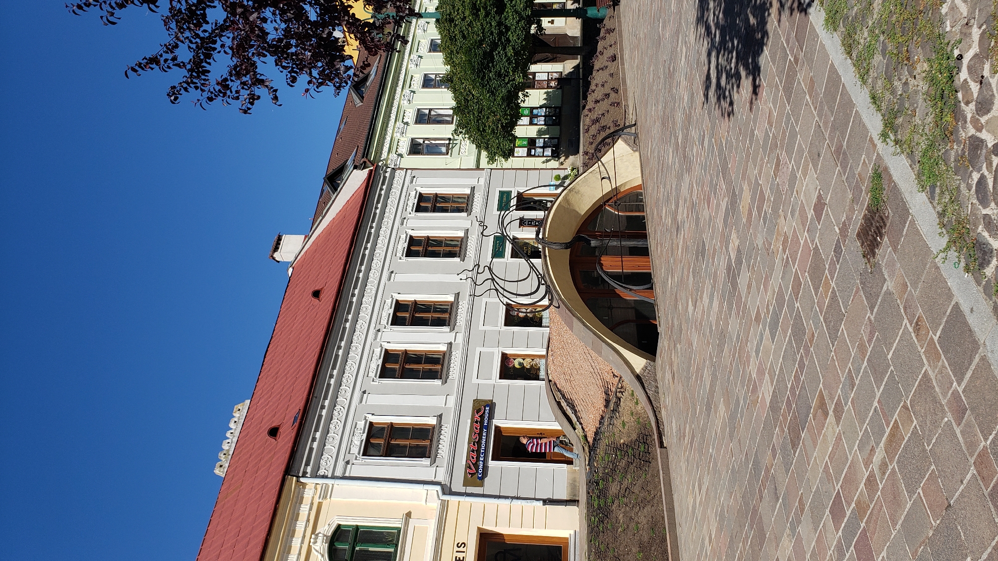 The image depicts the exterior of the confectionery house "Vatsak" located in Prešov, Slovakia. The building features a distinctive architectural element at its entrance: a subterranean passage with an arched roof covered in small, light brown tiles and surrounded by grass. A large, stylized metal sculpture resembling a snail is positioned atop the arched roof. The building itself is a multi-story structure with a red-tiled roof and light gray facade, featuring multiple windows with wooden frames. In front of the building is a paved plaza with square stone tiles in varying shades of brown and gray. A person wearing a striped shirt and jeans is walking out of the building's entrance. The sky is clear and blue. To the right of the building, a leafy tree provides shade.