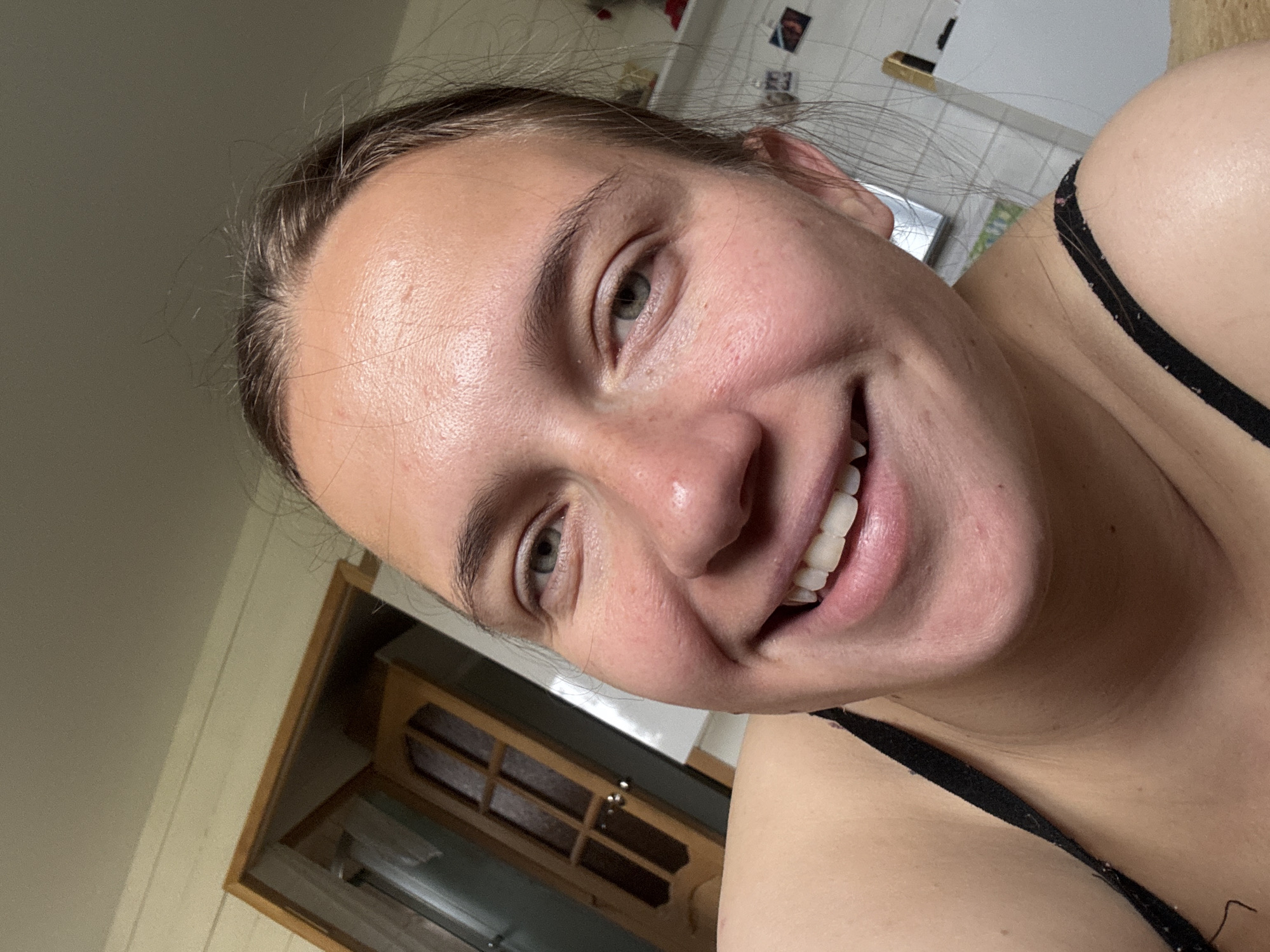 A close-up, slightly angled selfie shows a young woman smiling broadly. She has fair skin, light brown hair pulled back, and light-colored eyes. The image quality is slightly blurred. She wears a black tank top with thin straps. The background includes a wooden door with a glass panel, a white tiled wall, and a beige wall. It is impossible to confirm the location of the image, Раздолье, Russia, with the provided information.