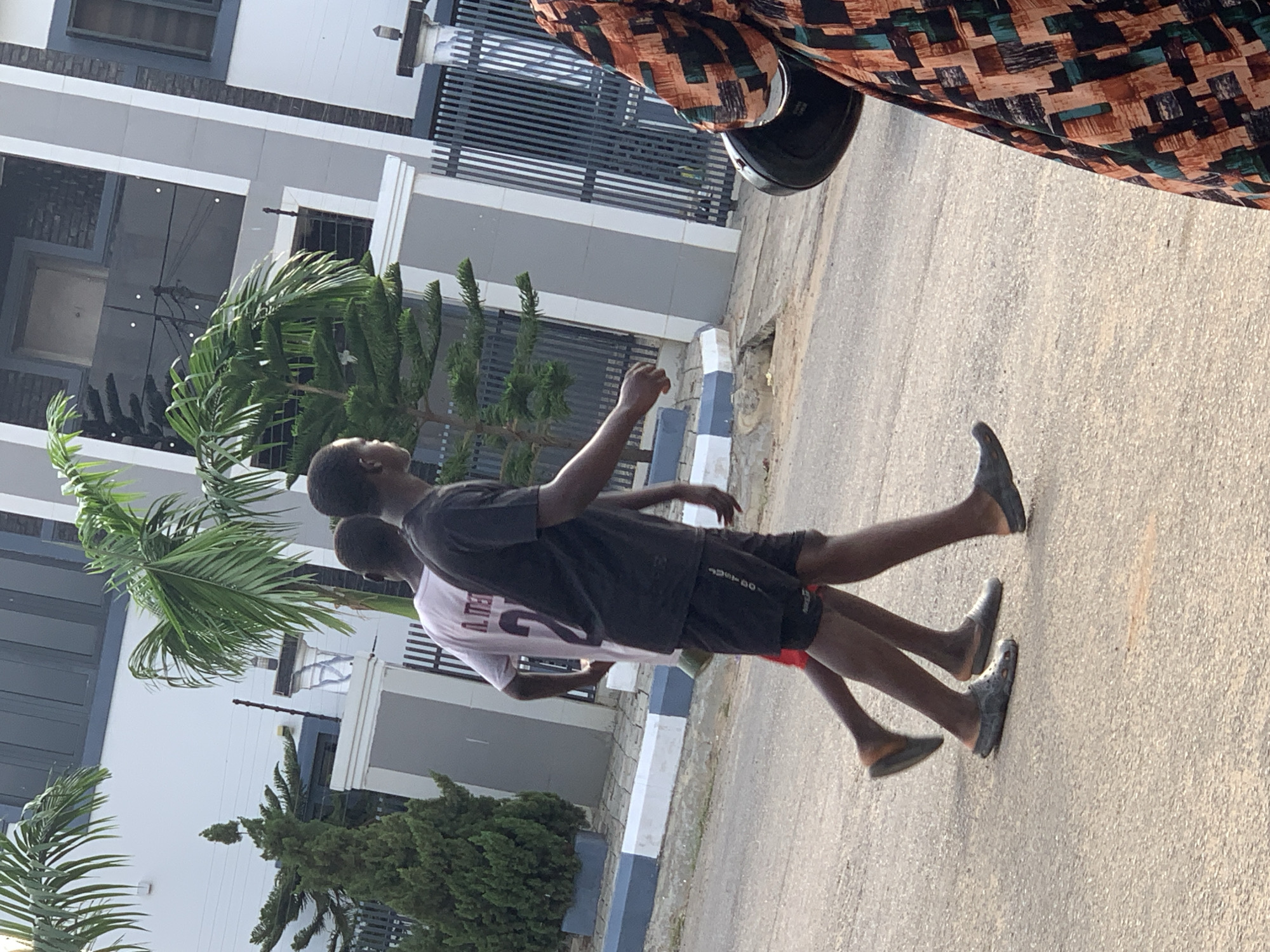 Two young men are walking on a paved road in Abuja, Nigeria. The man on the left wears a pink t-shirt with the number "27" and "A. MEN" printed on it, and dark shorts. He is wearing black clogs. The man on the right wears a black t-shirt and black shorts, and appears to be wearing grey sandals. In the background, a modern multi-story building with grey and white accents is visible. Several palm trees and a manicured evergreen tree are planted in front of the building. A black metal fence separates the property from the road. A person wearing a patterned dress and carrying a black helmet is partially visible on the right side of the frame. The lighting suggests it is daytime.