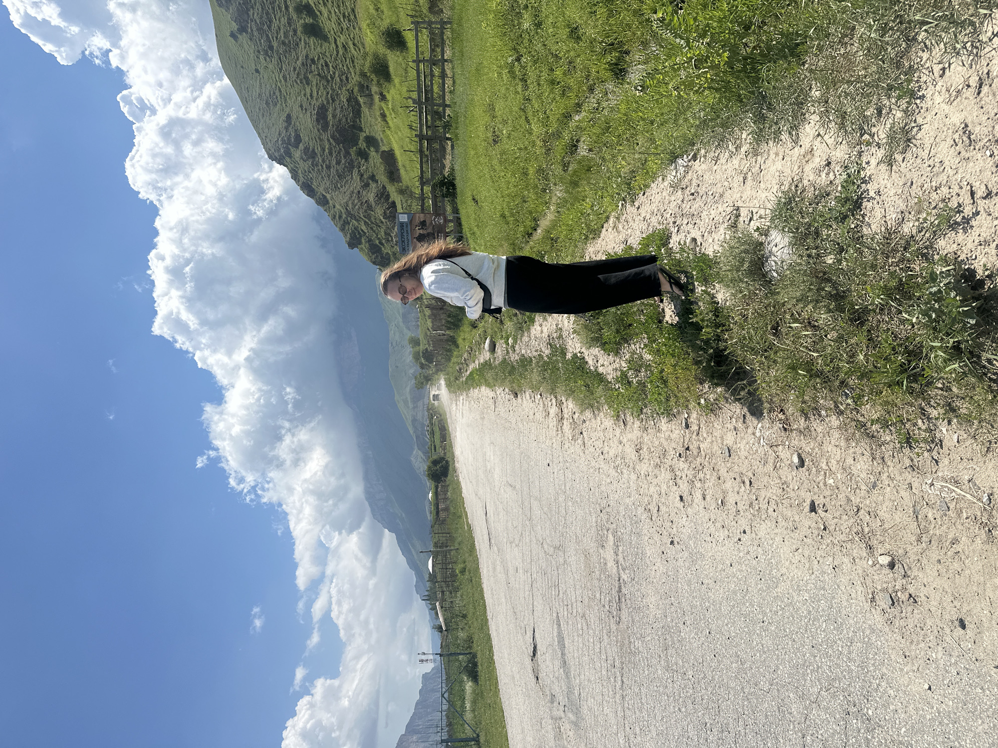 In Bylym, Russia, a young woman with long brown hair and sunglasses stands on a dirt path beside a cracked asphalt road. She wears a white crewneck sweatshirt and black pants. To her right, a green, grassy hillside rises, topped with a wooden fence and a sign. In the distance, mountains are shrouded in clouds, and a car can be seen on the road. The sky is a vibrant blue with large, puffy white clouds.