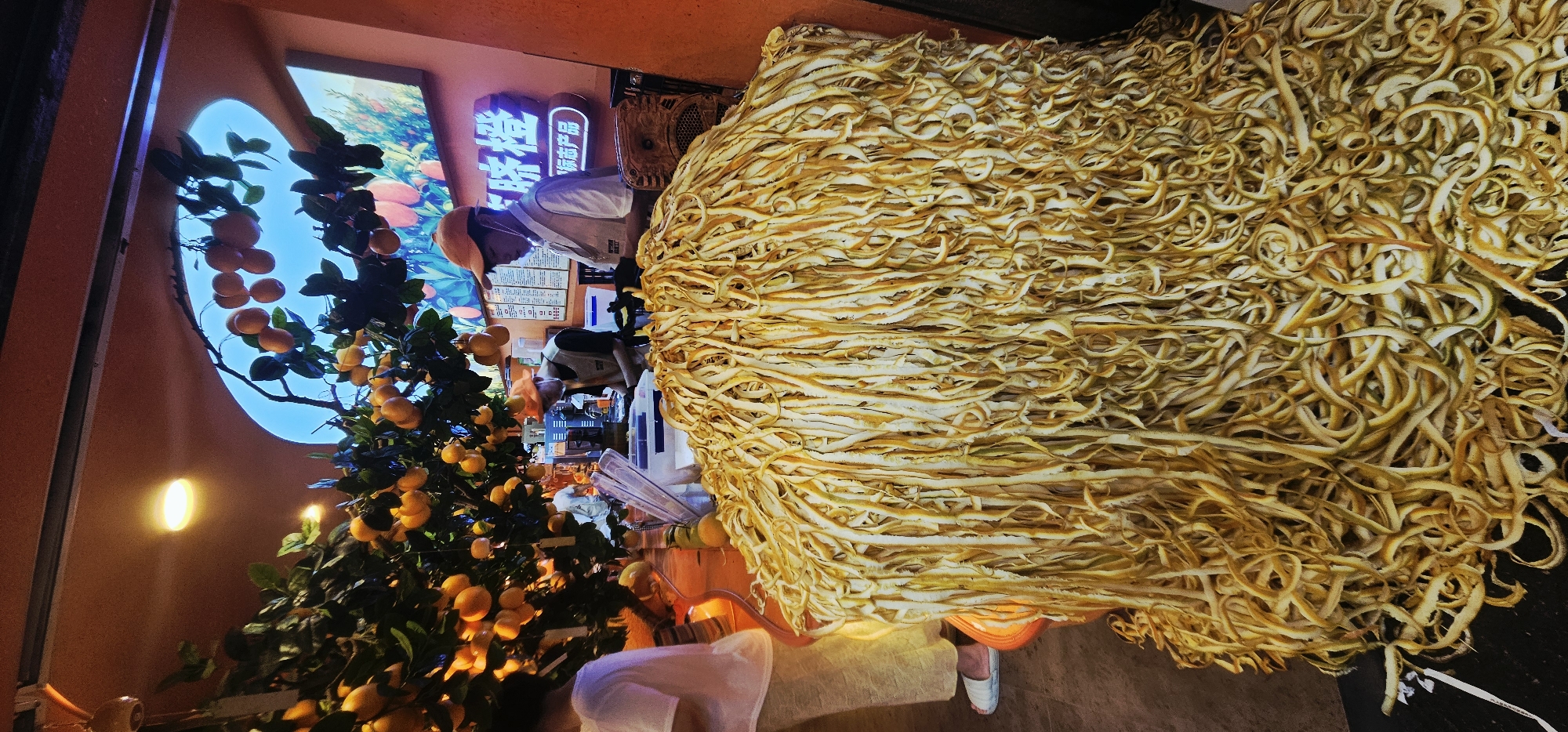 In Yuzhong District, China, a close-up shot captures a mountain of shredded citrus peels in the foreground, dominating the lower half of the frame. The peels, a mix of yellow and white, are long and stringy, piled high in an irregular mound. Behind this, a counter area is visible. To the left, a person in a white top and light-colored pants stands with their back to the camera, their feet clad in white sandals. To the right of the peel pile, a vendor wearing an orange cap and a light-colored vest over a white shirt is seen from the side, appearing to work at the counter. Another individual, partially visible behind the vendor, also seems to be working. Overhead, a large lemon tree, laden with ripe, orange-yellow fruit, extends branches into the scene, creating a canopy. The background features a window with a bright blue sky visible through an arched opening, and beyond that, a colorful mural depicting fruit and foliage. A neon sign with Chinese characters illuminates the upper right portion of the background. The overall lighting suggests an indoor setting with artificial light sources contributing to the warm, somewhat dimly lit atmosphere.