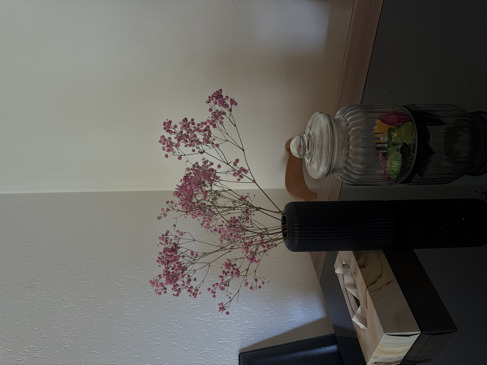 The media file presents an indoor still-life scene, likely in a home in Oberflachs, Switzerland. The primary subjects are decorative items arranged on a dark, reflective surface, possibly a table or console, against a light-colored, textured wall.
The central object is a bouquet of delicate, dried pink flowers, resembling Baby's Breath (Gypsophila), held in a tall, cylindrical black vase. The vase features a ribbed, matte finish and is positioned towards the left of the reflective table. The small pink blossoms spread gracefully, creating a soft contrast against the light wall.
To the right of the floral arrangement, a clear, fluted glass jar with a domed lid rests on the table. The jar contains colorful items, appearing to be individually wrapped candies or small treats. Further to the right and slightly below the jar, a rectangular tissue box is visible. This tissue box has a light-colored pattern resembling marble or a light wood grain with subtle brown veining.
The dark, polished surface of the table reflects ambient light and portions of the objects, adding depth to the composition. In the background, behind the table on the right side, a narrow vertical section of a brown or wooden panel is visible, suggesting part of a cabinet or shelving unit. A dark, possibly upholstered, surface can be seen at the bottom right corner, indicating a nearby piece of furniture like a chair or sofa.
The lighting appears soft and even, typical of an indoor environment, creating subtle shadows and highlights. No specific activity is depicted, and no text is visible within the frame. The overall scene conveys a calm and decorative domestic setting.