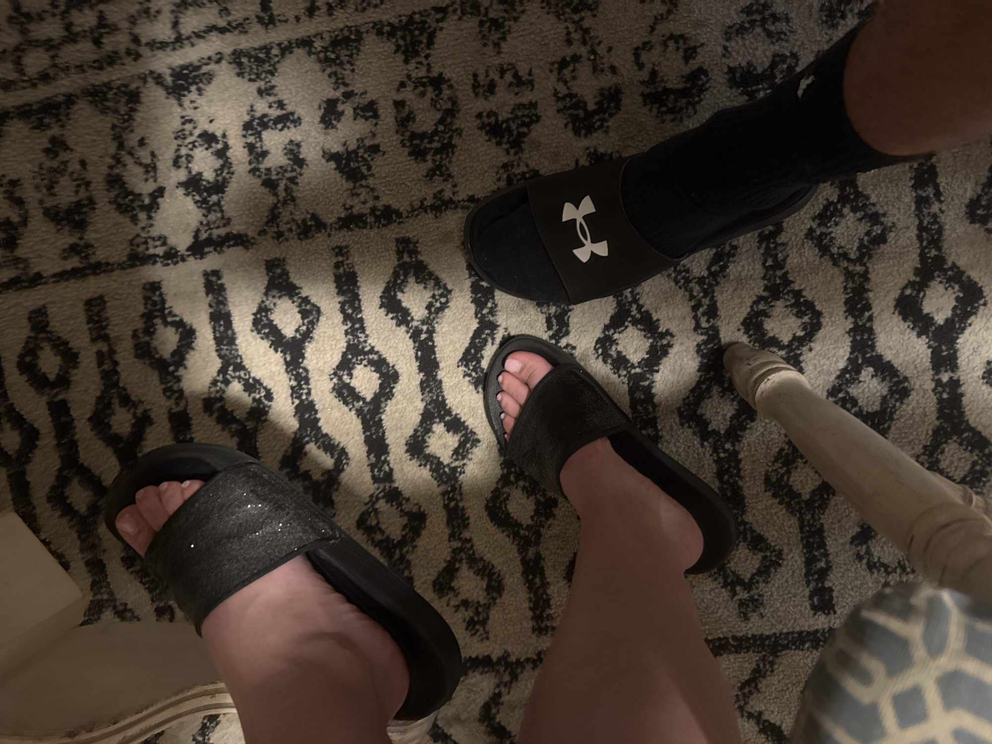 The image shows a close-up of two people's feet resting on a patterned rug. One person is wearing black slide sandals with glitter detailing on the strap, and the other is wearing black slide sandals with the Under Armour logo and black socks. The rug has a cream background with a black abstract geometric pattern. The lighting is dim, suggesting an indoor setting. The image is undated and unlocated, and no specific city or country is mentioned.