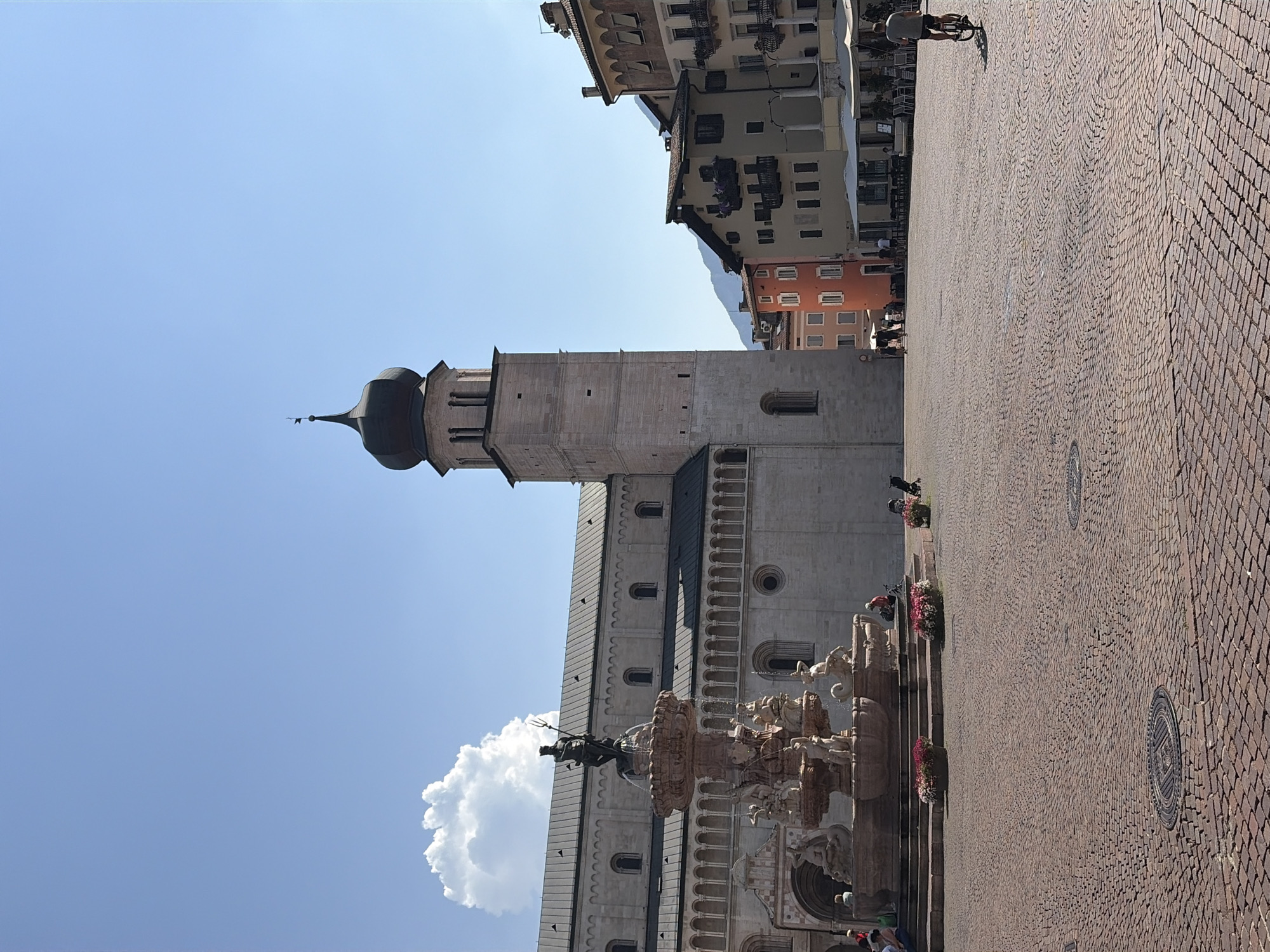This is a wide-angle, eye-level shot of Piazza Duomo in Trento, Italy, on a clear, sunny day. In the foreground, a vast expanse of cobblestone pavement is visible, with manhole covers embedded in it. To the left, a large, ornate fountain features multiple tiers and numerous sculptures, including a central figure of Neptune holding a trident. Behind the fountain, the substantial facade of the Trento Cathedral (Duomo di Trento) is prominent, characterized by its grey stone construction and long, arched windows. Adjacent to the cathedral is a tall, square bell tower with a dark, domed roof.
The middle ground reveals a street scene with several colorful buildings housing shops and cafes. These buildings vary in height and architectural style, with balconies adorned with flowers. People are scattered throughout the square: some are seated near the fountain, others walk, and one person is riding a bicycle away from the camera towards the right side of the frame. Umbrellas of outdoor seating areas are visible in front of some establishments.
In the background, distant mountains can be seen through a gap between the buildings. The sky is a bright, clear blue with a single, large cumulus cloud to the left of the bell tower. The overall atmosphere is one of a bustling city square on a pleasant day.