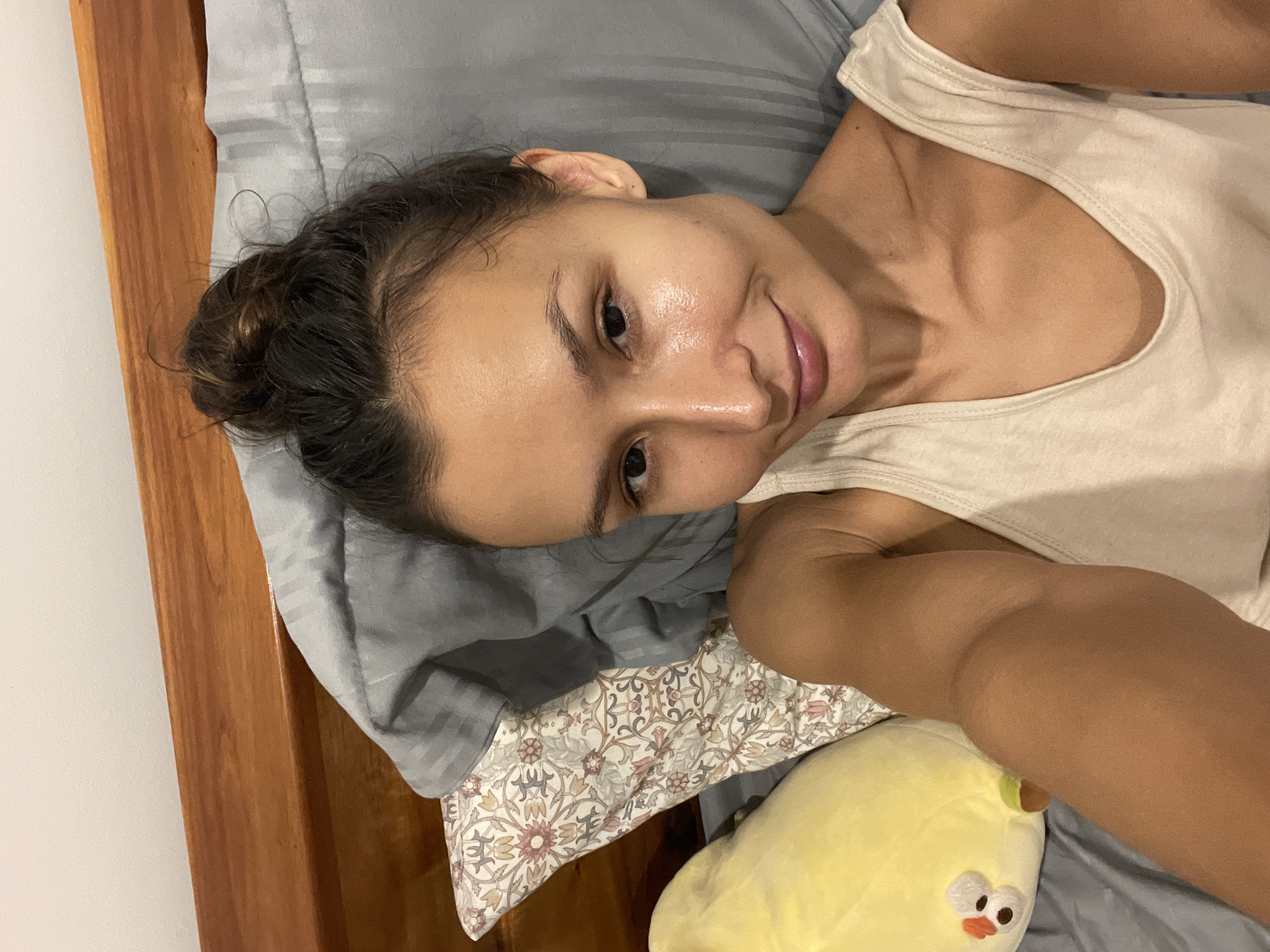A young woman is captured in a close-up, upward-angled selfie, lying on her side in what appears to be a bed in Mũi Né, Vietnam. She is looking directly at the camera with a soft, natural smile, her dark eyes open. Her dark hair is pulled back into a casual bun, with some wisps around her forehead. Her skin appears bare and has a natural sheen. She is wearing a light-colored, possibly off-white or beige, sleeveless tank top.
The setting is an indoor bedroom. Her head rests on a grey pillow with subtle stripes, and further down the bed, there is a pillow with a light-colored floral or botanical pattern in shades of pink, grey, and cream. A wooden bed frame is visible on the left side of the image, adding a warm, rustic touch to the scene. Below her right arm, a bright yellow plush toy resembling a chick or duckling with an orange beak and cartoonish eyes is visible, resting on a grey sheet. The overall impression is one of relaxation and comfort, likely captured during a casual moment indoors. The lighting is soft, suggesting an ambient indoor environment. There is no visible text in the image.