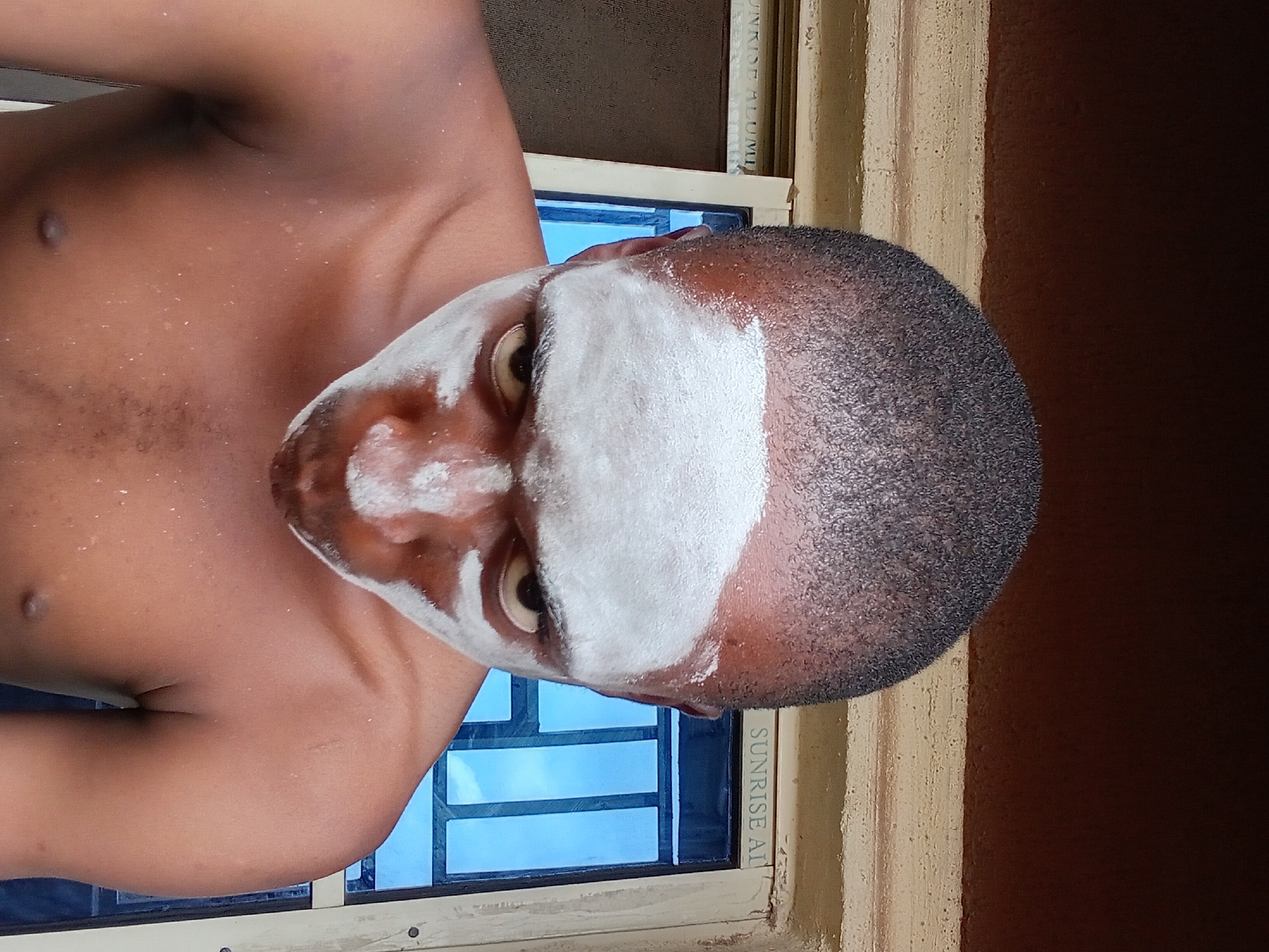 A close-up, eye-level shot captures a dark-skinned person in Nkpor, Nigeria, with white powder applied to their face and forehead. The person is shirtless, revealing their shoulders and chest, and is looking down towards the camera. They have dark, short-cropped hair. Behind them is a window with blue panes and a white frame, with what appears to be a brown wall above. The lighting suggests it is daytime.