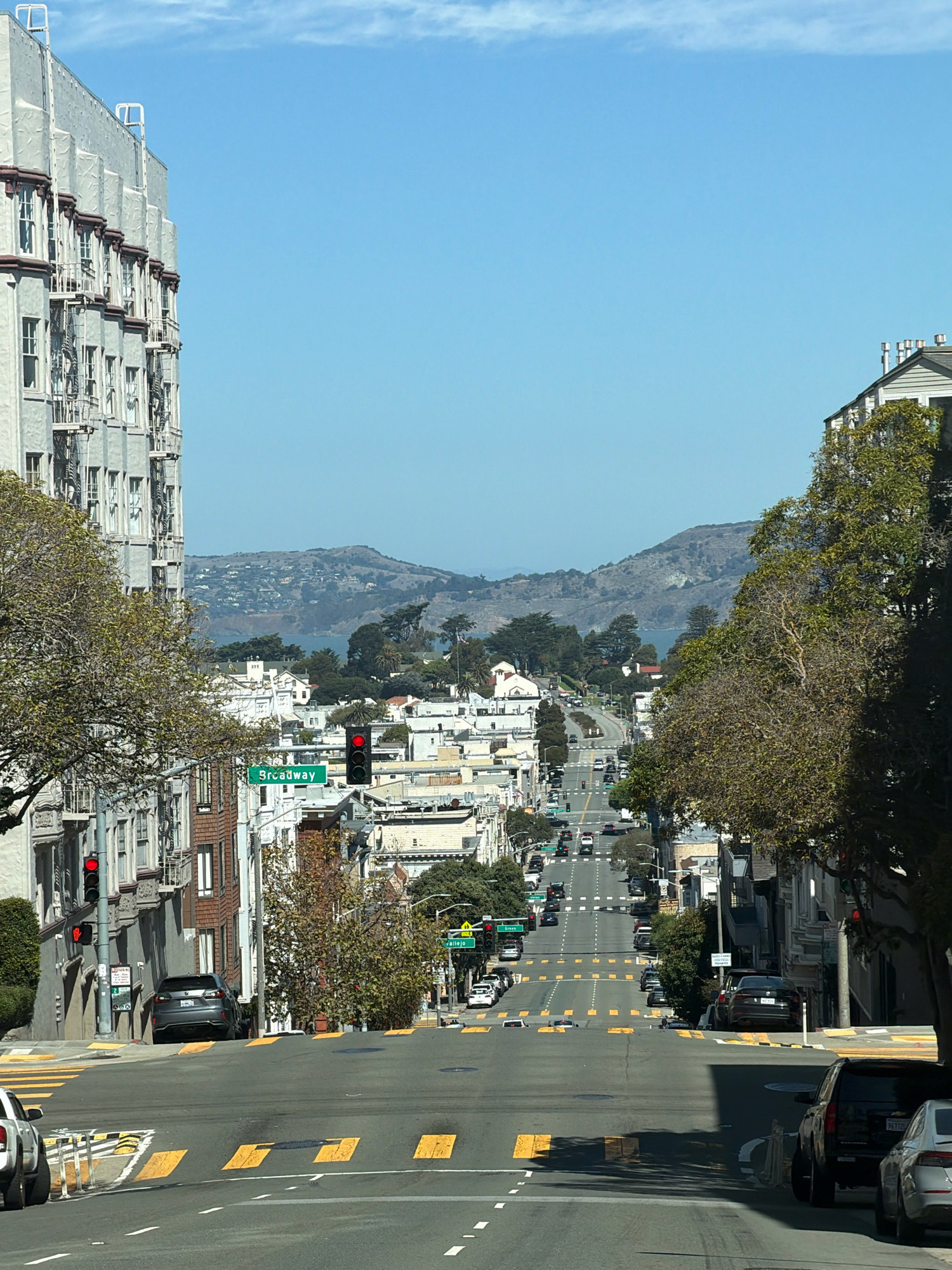 The image depicts a view looking down a steep, wide street in San Francisco, California, USA. The street is lined with multi-story buildings, many with architectural details characteristic of the city, such as ornate facades and fire escapes. Trees with sparse foliage line the sidewalks. The street is paved with asphalt and features yellow crosswalk markings and speed bumps. Several cars are visible parked along the street and in motion, navigating the incline.
In the mid-ground, a street sign indicates "Broadway." Traffic lights are present at intersections, with one red light clearly visible. In the distance, the street opens up to a view of the San Francisco Bay with rolling hills and structures visible on the opposite shore. The sky is a clear, bright blue with a few wispy clouds. The overall impression is of a typical San Francisco street scene on a sunny day, characterized by its hilly terrain and dense urban architecture.