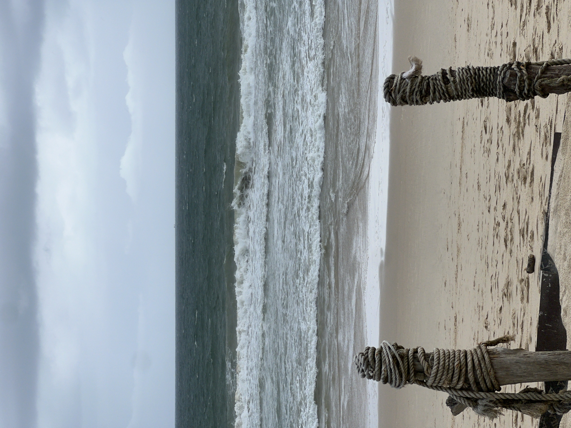 A seascape features a beach on the right side, with two wooden posts wrapped in rope partially visible in the foreground. The beach transitions into foamy waves of the ocean, and further out, the darker expanse of the sea stretches to the left side of the frame. A cloudy sky is visible above the sea. The location is unknown.