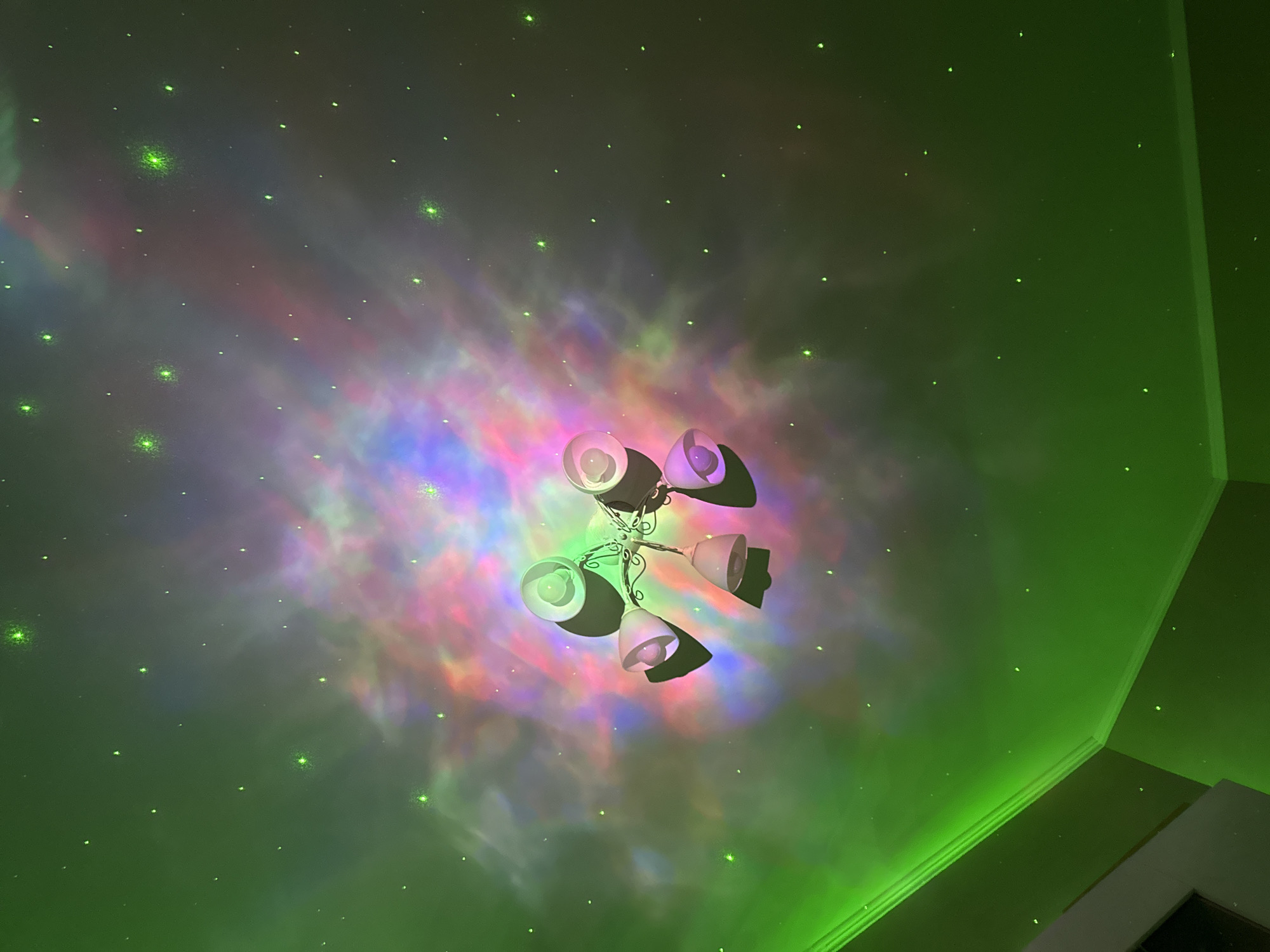 The image captures a view looking upwards towards a ceiling with a chandelier fixture in the center. The ceiling is illuminated with a projection creating the effect of a nebula or colorful cosmic clouds with specks resembling stars. The walls are visible on the right side of the image and seem to be illuminated with a green light. The location appears to be an interior space, but the exact location, including the city, is unknown.