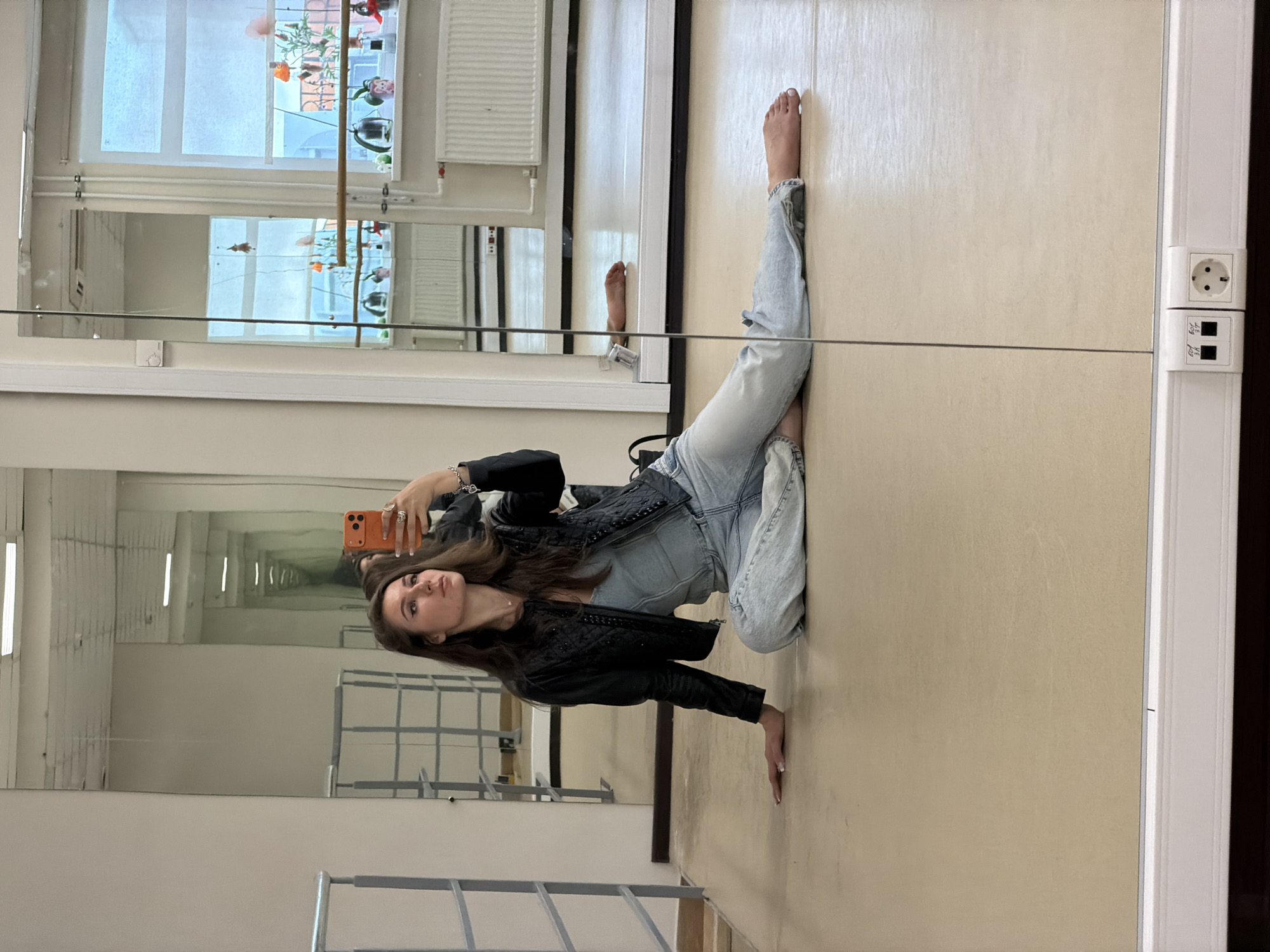 A young woman with long, dark hair is sitting on the floor of what appears to be a dance studio or rehearsal space, taking a selfie in a large mirror. She is wearing a denim corset-like top, a black quilted jacket, and light-wash baggy jeans. Her left arm is resting on the floor, and her right hand is holding an orange smartphone, angled to capture her reflection. Her gaze is directed towards the phone screen. The studio floor is a light, neutral color, and a large mirror occupies a significant portion of the background. Reflections in the mirror show rows of lockers or shelves, a barre across a window with a view of trees and buildings, and what appears to be an electrical outlet on the wall at the bottom of the frame. There are no other people or animals visible in the immediate vicinity. The setting is indoors, with ample natural light coming from the windows.