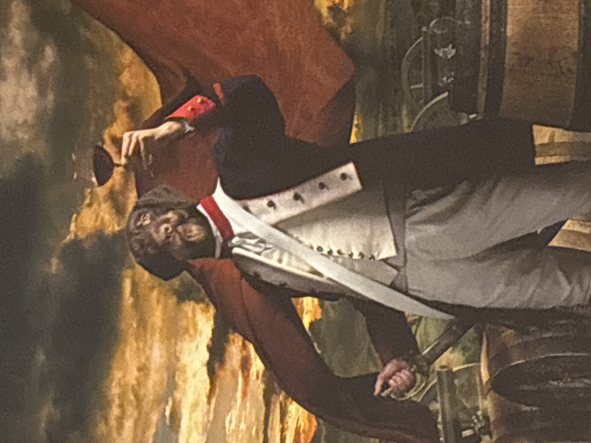 The image is a portrait of a man, possibly from the 18th or 19th century, judging by his clothing. He wears a black coat with red trim on the sleeves and a white waistcoat. His pants are light gray or white. A large red cloth or cape drapes behind him. His hands appear to be tied together. The background appears to be an outdoor scene with a mix of light and dark areas. The location is unknown.