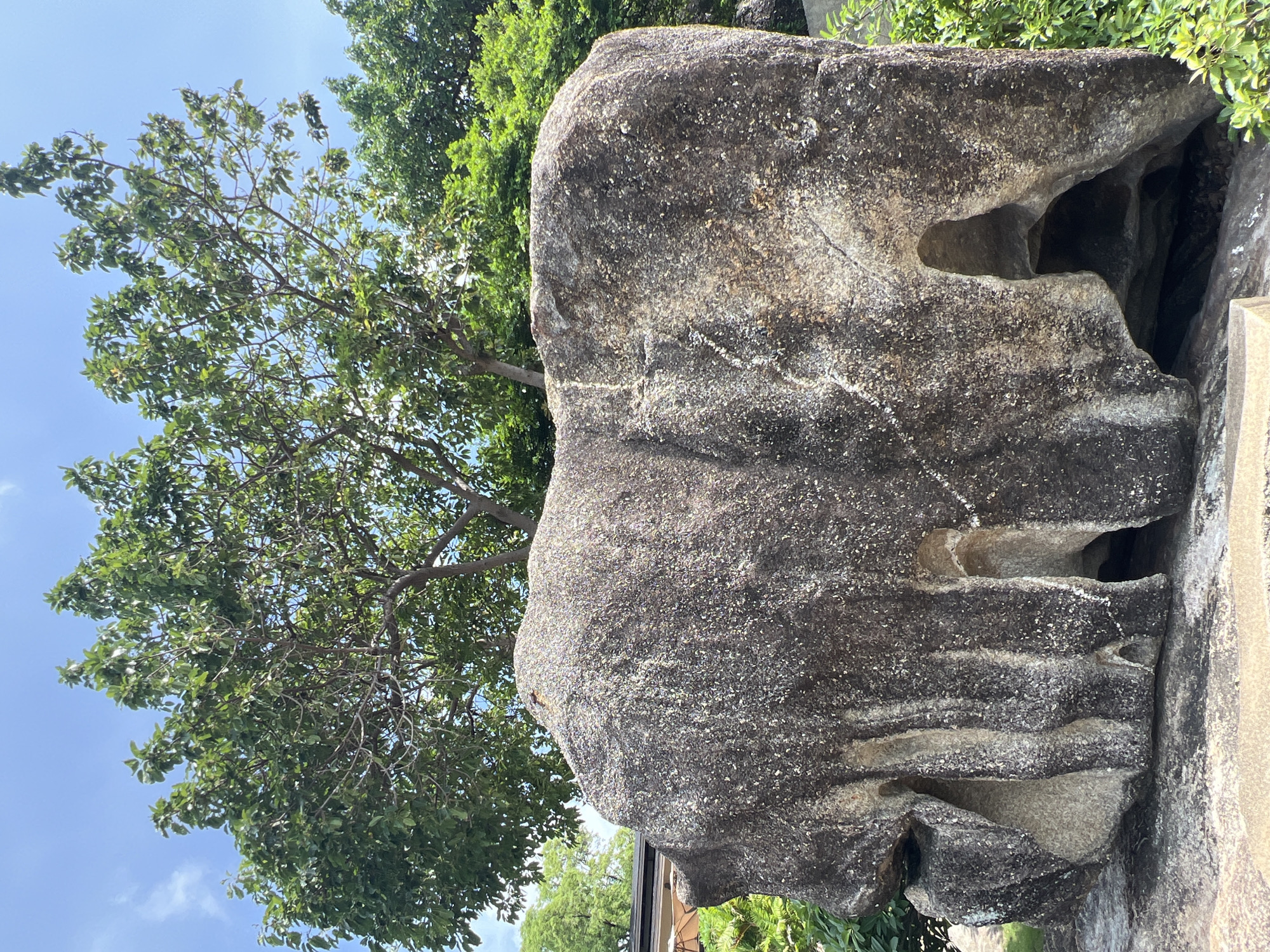 The image shows a large, elephant-shaped boulder in a park-like setting with lush green trees and a bright blue sky. The boulder is grey with darker veins and appears to have been naturally eroded into the shape of an elephant. There are openings in the rock that resemble legs or crevices. The boulder is situated in what appears to be a public space, with hints of man-made structures and umbrellas visible in the background, suggesting a tourist attraction or a landscaped park. The overall impression is of a natural wonder that has been integrated into a recreational area. The location is identified as Baan Chaweng Noi, Thailand.