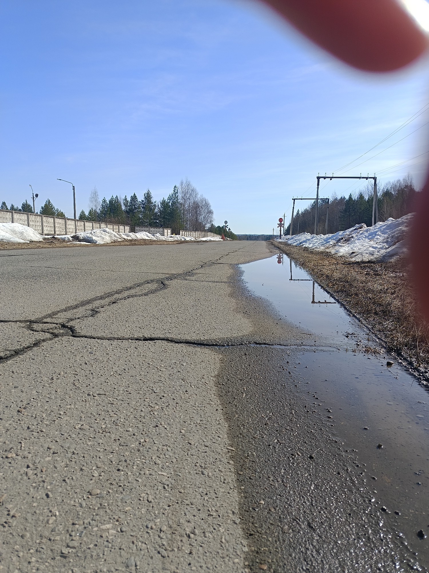 The image depicts a low-angle view of an asphalt road in Chaikovsky, Russia. The road shows significant cracking. A large puddle of water occupies the right edge of the road. Patches of snow remain on either side of the road, near fences and trees. A stop sign is visible in the distance. Overhead power lines and utility poles are also present. A blurred finger partially obscures the upper right portion of the image. The sky is clear and blue. The overall environment suggests early spring with melting snow.