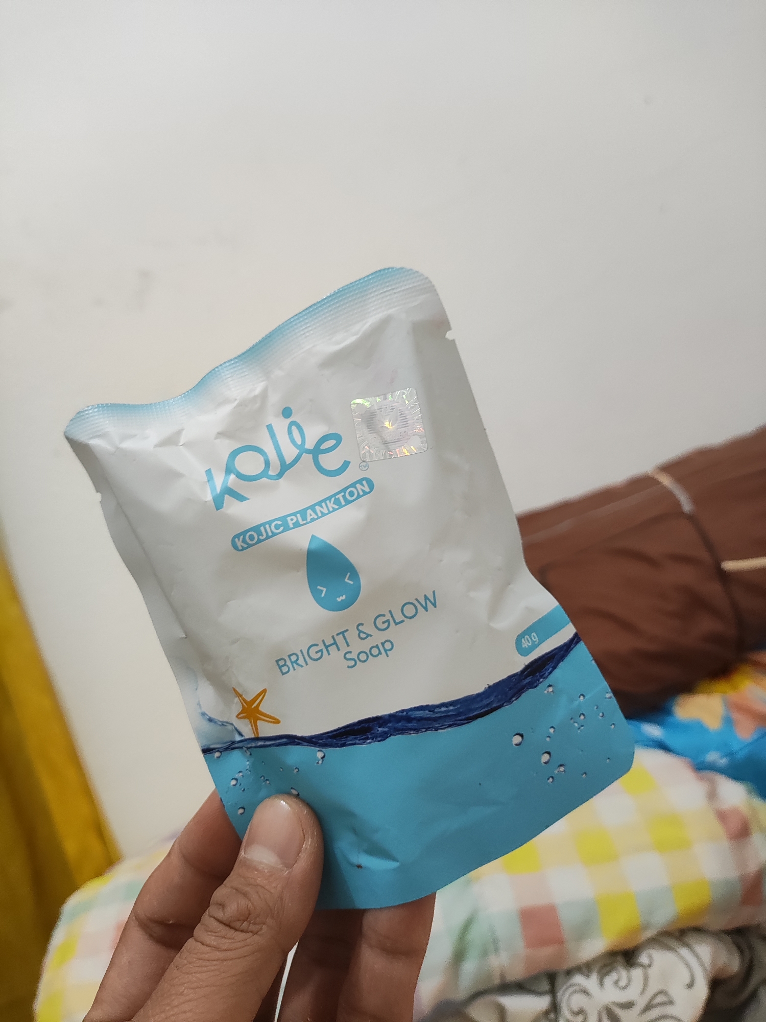 A person's hand holds a small, white and light blue packet of Kojie-san soap in the city of Wanajaya, Indonesia. The packet features the brand logo "Kojie" in blue cursive script, with text indicating "KOJIC PLANKTON" and "BRIGHT & GLOW Soap". A stylized blue water droplet with a smiley face is printed on the packet, along with a starfish and water bubbles. The background consists of a white wall, a brown blanket, and a colorful checkered textile. The lighting suggests an indoor setting.