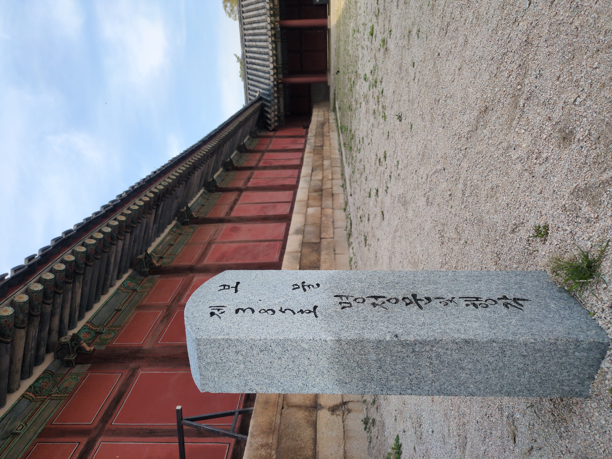 The image depicts a low-angle, side view of a stone marker in Seoul, South Korea. The marker, light gray with dark writing, sits at the edge of a gravel pathway. Behind the marker is the red and brown wall of a traditional Korean building, topped with an ornate roof structure of dark gray tiles. The sky, visible above the building, is a light blue with faint clouds. The gravel path is light brown and sparsely covered with small patches of green vegetation.