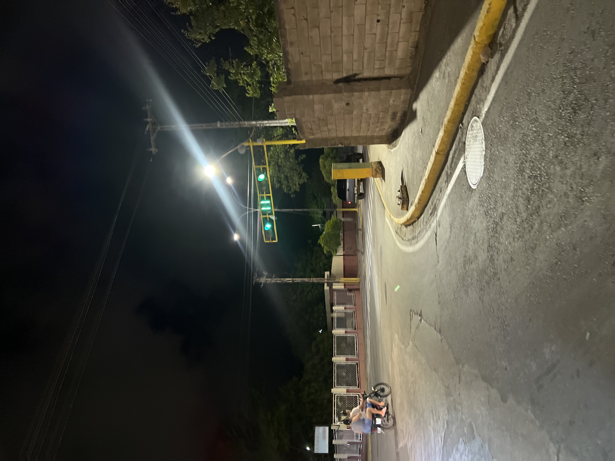 The image depicts a street scene at night in Juan Griego, Venezuela. A motorcycle with two occupants, a male driver and a female passenger, is traveling away from the viewer on the left side of the frame. The motorcycle is moving across a road intersection marked with white pedestrian crossing lines. In the background, there are buildings with chain-link fences and some trees. A traffic light with green signals is illuminated and suspended over the intersection by a yellow metal structure. To the right, a brick wall borders the street, and a small, dark-colored SUV is visible further down the road. The street is paved, with a yellow-painted curb running along the right side. A manhole cover is present on the road surface. The sky is dark, with some streetlights and power lines visible.
