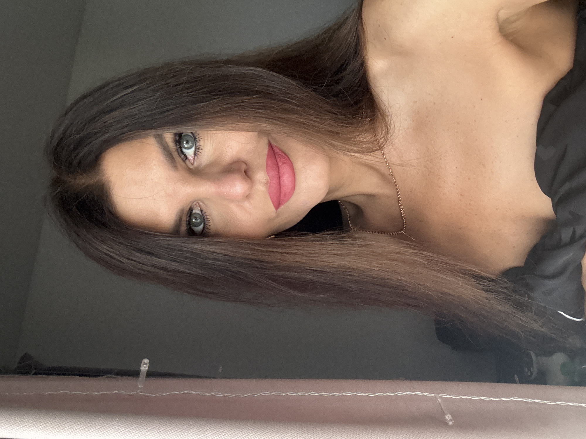 A middle-aged woman with long, dark brown hair and blue eyes is pictured in a close-up, front-facing shot. She is wearing pink lipstick and a delicate gold necklace. Her skin appears fair with some freckles visible on her shoulders and décolletage. She is wearing a strapless black garment. To the left of the frame, a sheer pink curtain hangs, with visible stitching and a small, clear plastic clip. The background is a plain gray wall. The lighting suggests the photograph was taken indoors, likely near a window due to the curtain. The image does not contain any specific geographical markers that would indicate its location in Chaikovsky, Russia.