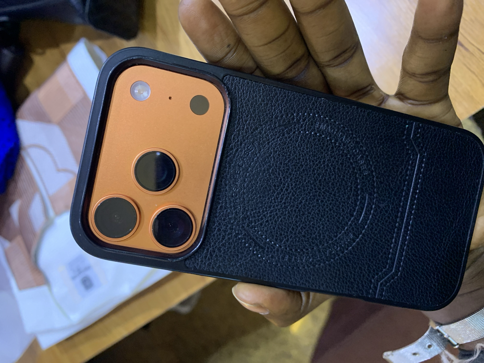 A person's hand is holding a black iPhone case with an orange camera bump. The camera bump has three lenses and a flash. The case is made of textured leather and has decorative stitching. In the background, a white garment with brown stripes is visible, along with a wooden surface. The scene appears to be indoors, possibly on a desk or table. There is no information about the location in Gura Top, Nigeria.