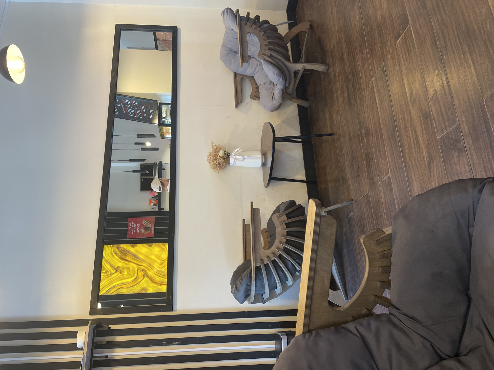 The image depicts the interior of a cafe or lounge area. Two uniquely designed wooden chairs with dark grey cushions are present. One chair is in the foreground, partially obscured, and faces left. The other chair is positioned to the right of a small, dark, triangular side table, which holds a white vase with dried flowers. A large, wide mirror hangs on the wall above the chairs, reflecting the interior and a menu board with text that appears to be in Cyrillic. The floor is made of dark wood planks. The cafe is likely located in Radkevshchina, Russia, as indicated by the menu's script.