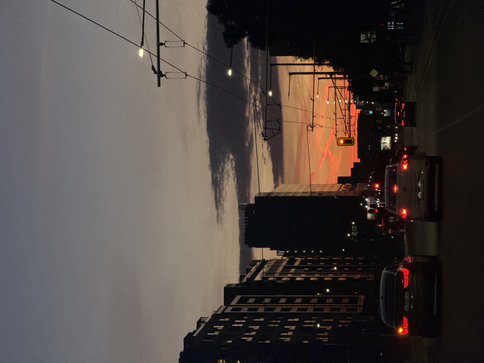 The image depicts a street scene at dusk in Sofia, Bulgaria. Buildings line the left side of the frame, and a row of cars sit on the right side of the frame. The sky features a mix of dark gray and orange hues, likely from a setting sun. Overhead tram lines stretch across the sky. A traffic light hangs over the street, displaying a red signal. Vehicle tail lights are illuminated, indicating stopped traffic. The scene is urban and suggests a busy time of day.