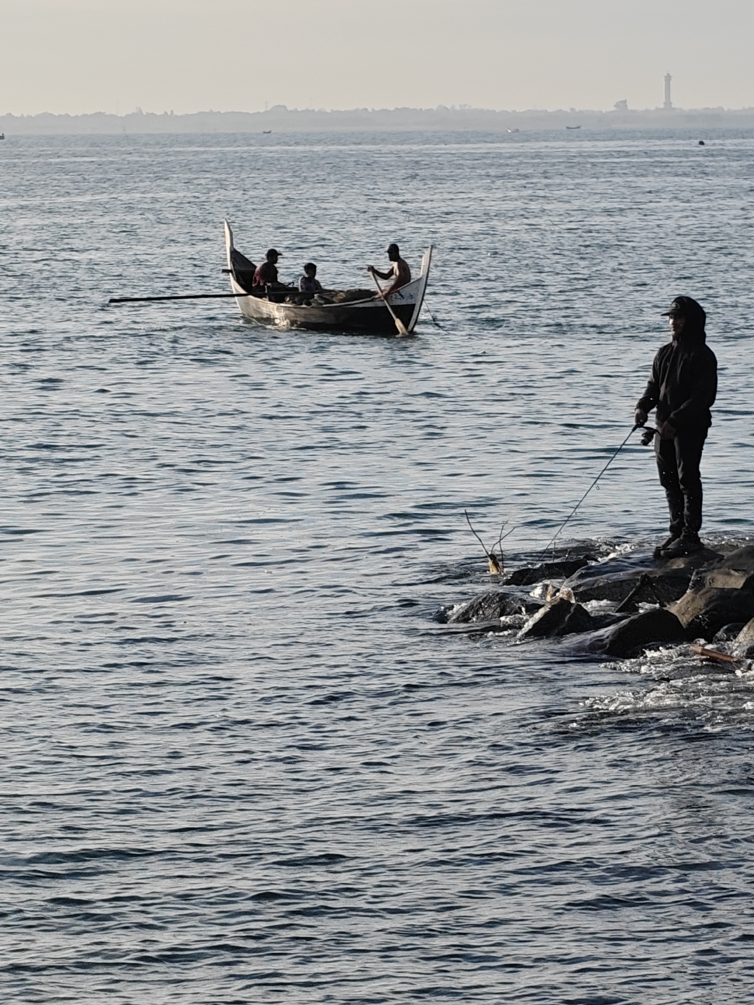 The image shows a coastal scene in Lhokseumawe, Indonesia. In the foreground, a man dressed in dark clothing and a cap is standing on a rocky shoreline, holding a fishing rod. The sea is visible in the middle ground, with gentle waves. A small wooden boat with three people aboard is navigating the water. In the distance, a hazy horizon with a faint outline of a lighthouse and other structures can be observed.