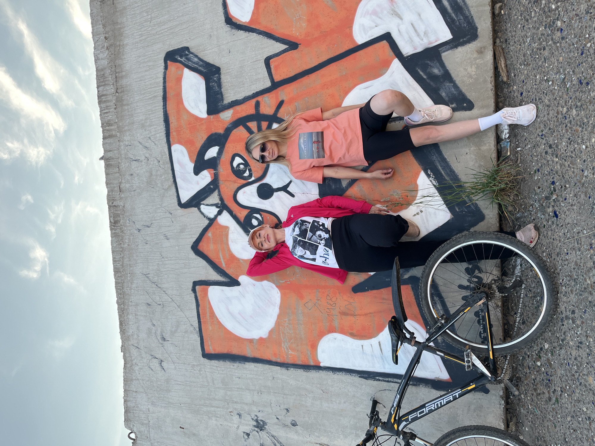 Two women pose for a photograph in front of a graffiti-covered concrete wall in Chaikovsky, Russia. The graffiti depicts an orange and white abstract design with what appears to be a cat's face. A black bicycle with yellow accents is parked to the left of the women. The ground is covered in gravel and small debris. The sky above is blue with scattered clouds. The woman on the left is wearing a pink jacket over a white t-shirt and black pants, with a beige baseball cap. The woman on the right is wearing a coral-colored t-shirt, black biker shorts, and sunglasses.