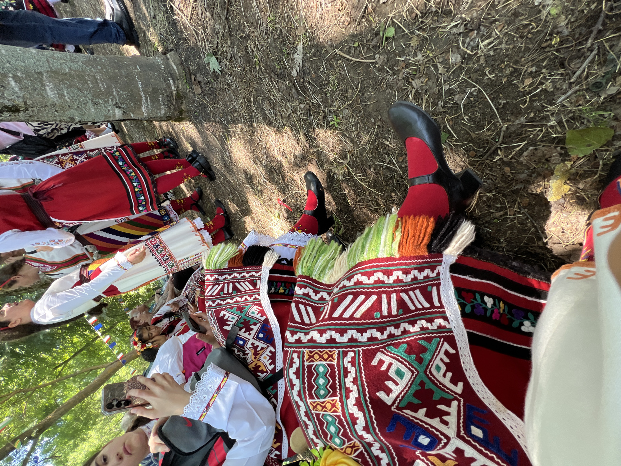 This image depicts an outdoor scene in Bolyarovo, Bulgaria, featuring multiple individuals in traditional Bulgarian folk costumes. In the immediate foreground on the left, a young woman is seated, holding and looking at a smartphone with a gold/rose gold frame and a three-camera array. She wears a white long-sleeved blouse with lace cuffs and red/yellow trim, a dark vest, a red apron-style garment with intricate geometric and floral patterns in white, green, blue, yellow, red, and black, and has vibrant green and orange fringed tassels extending from her costume. Her leg is visible, showing a red stocking and a black Mary Jane-style shoe with a strap and heel. A gray Nike backpack rests next to her.
To her right, partially obscured, another person's foot is visible, also wearing a red stocking and a black shoe. Further back, several other individuals are seated and standing. Another young woman with a floral headpiece (red and yellow flowers) is seated, also looking at a smartphone. An older woman in a white blouse and dark attire is seated behind her.
In the middle ground, several women stand wearing variations of traditional costumes, characterized by white blouses, colorful striped or red skirts/aprons with embroidered bands at the hem, often featuring floral or geometric motifs. Red stockings and black Mary Jane-style shoes are common footwear. One woman wears a white blouse, a red apron with multi-colored embroidery, and a vertically striped skirt. Another has a white blouse and a vibrant striped skirt.
On the far right, a large tree trunk with rough bark is prominent. Behind it, the lower legs of a man wearing blue jeans and black dress shoes are visible.
The ground consists of dry earth, scattered dry leaves, twigs, and sparse patches of green vegetation, indicating an outdoor, possibly shaded, environment. Overhead, the canopy of trees is visible. In the background, strung between trees, a line of colorful rectangular bunting flags (red, white, blue, green, yellow) suggests a festive event. The scene is illuminated by dappled sunlight, creating areas of light and shadow on the ground.