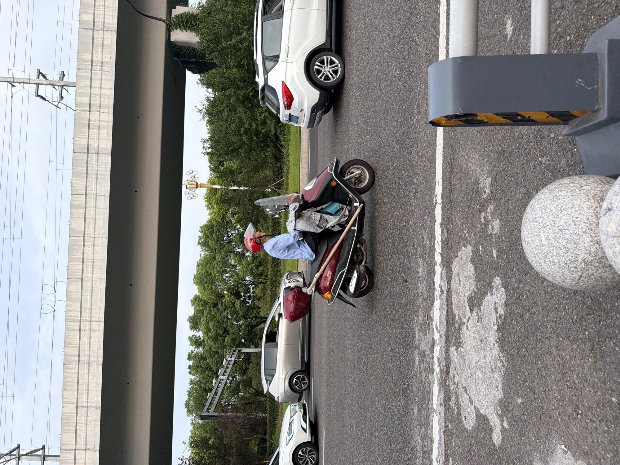 The image captures a street scene in Jiangjin, China, under an overcast sky. A person wearing a red helmet and a blue shirt is riding a red and black scooter. The scooter has a windshield and a red cargo box attached to the back. The rider is carrying a light blue bag on the handlebars. The scooter is on a paved road with a white line marking a lane. To the left of the scooter, a silver car is visible in an adjacent lane, and a white car is parked further ahead. To the right of the scooter, a white SUV is also on the road. In the background, a large concrete overpass dominates the upper portion of the image, with power lines running overhead. Trees and greenery are visible beyond the road, and a lamppost with multiple lights stands on the left side. In the foreground, the edge of a road barrier with yellow and black hazard stripes is visible, along with large spherical decorative elements.