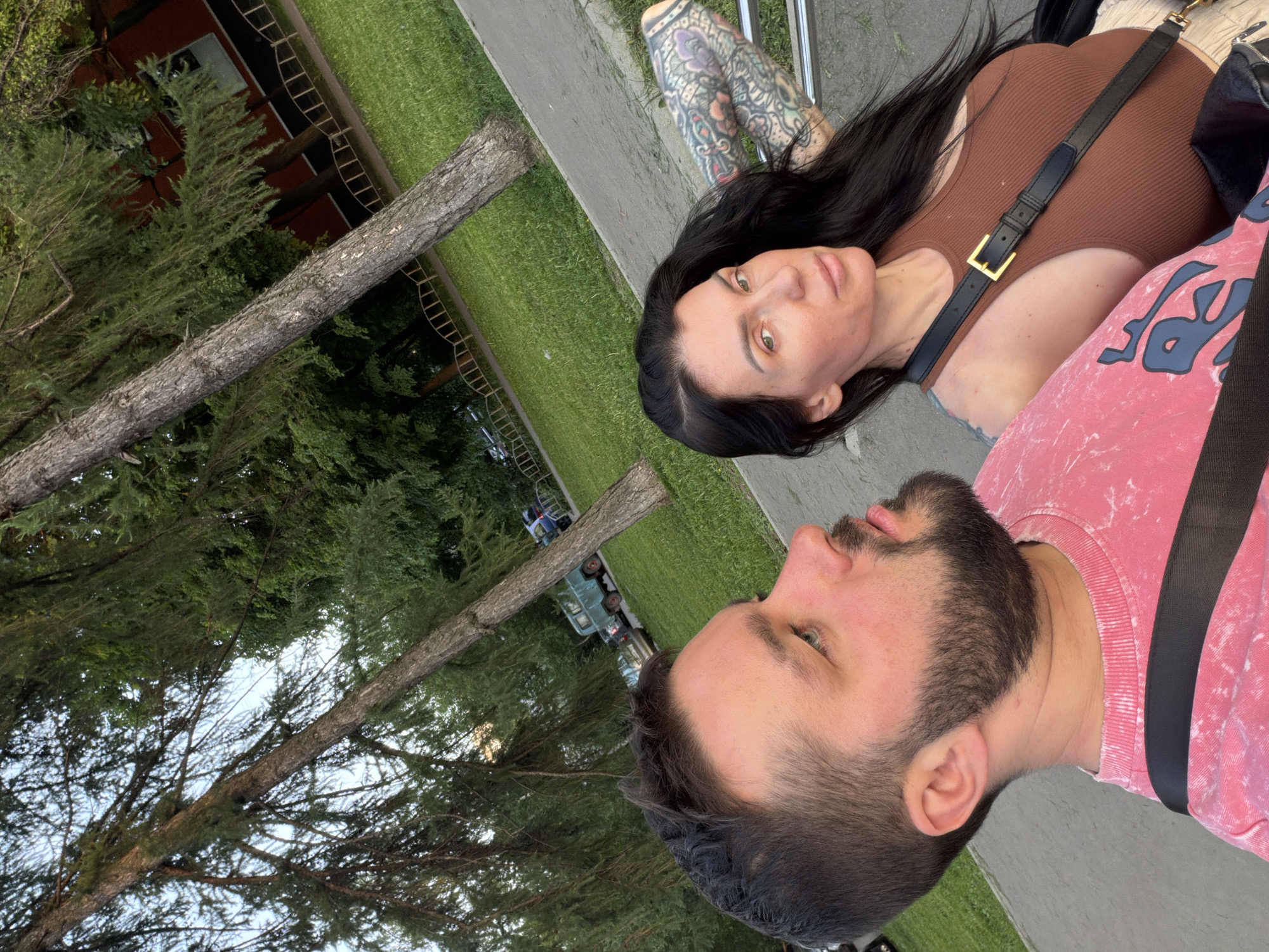 A photo depicts a man and a woman outdoors in Moscow, Russia. The man, on the right, has a beard and is wearing a pink shirt. The woman has dark hair and is wearing a brown top with a black purse strap. She has tattoos on her left arm. They are situated next to a sidewalk with green grass and trees in the background. An apartment building is visible in the distance.