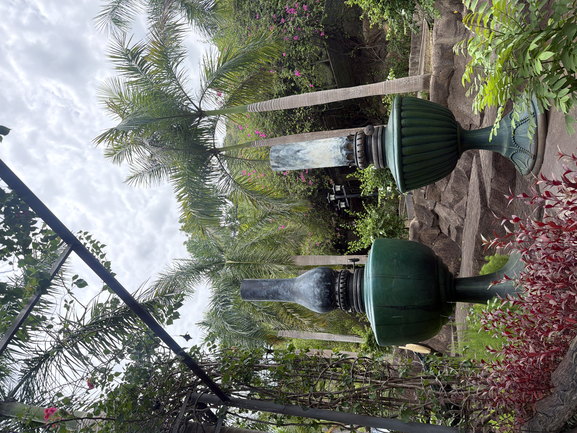 The image shows two vintage oil lamps standing amidst lush greenery and palm trees, suggesting an outdoor setting. The lamps are large and ornate, one made of dark metal with a flared base, and the other a lighter, weathered metal with a more cylindrical shade. Both are set on decorative pedestals. The background features a dense arrangement of palm trees and flowering shrubs, with a cloudy sky overhead. Rocks and paved pathways are visible in the midground and background, indicating a landscaped area. This scene is located at the Ocean Window Resort in Nha Trang, Vietnam, a resort known for its beautiful gardens and tranquil atmosphere. No people are present in the image.