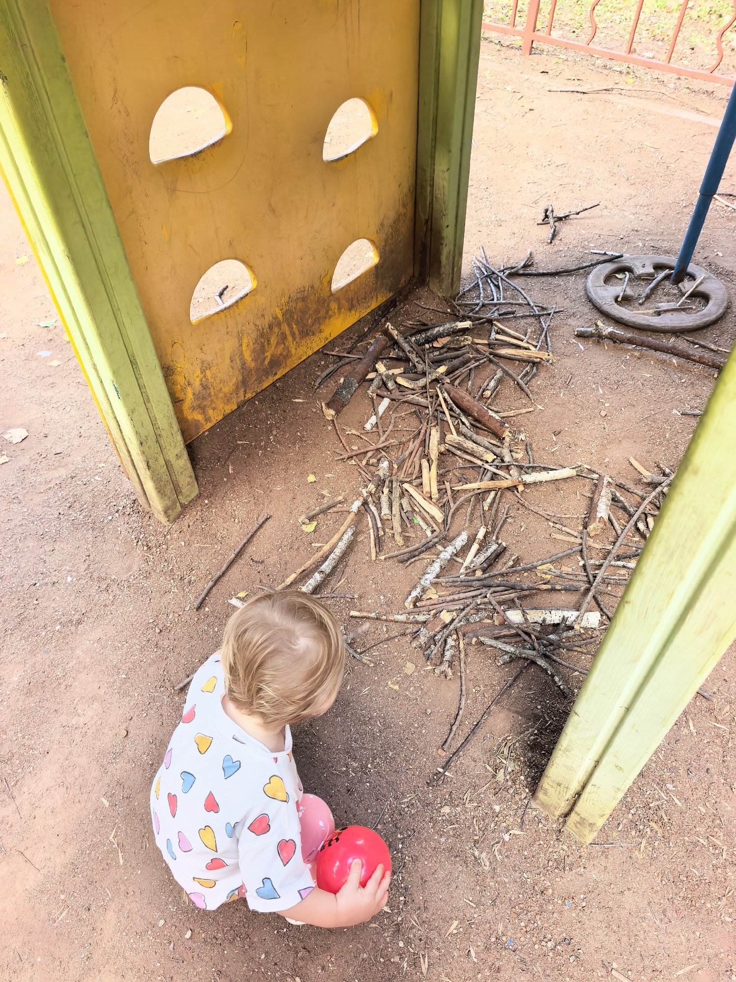 A blonde-haired child, facing away from the camera, sits on the ground. The child is wearing a white t-shirt with colorful heart patterns. In their right hand, the child holds a red, spherical object, possibly a ball. The ground appears to be made of reddish-brown dirt and small pebbles. To the child's right, a large pile of dry, broken sticks and branches is scattered. Behind the child and to the left, a yellow, modular playground structure is visible, with green frames. The structure has several semi-circular openings on its yellow surface. To the right of the pile of sticks, a dark, circular object, likely a weight or base, is partially obscured by sticks and a blue pole. In the background, a reddish-brown metal fence is visible, with greenery beyond. The overall setting suggests an outdoor playground in Sterlitamak, Russia, where the child is interacting with the environment and playing with a ball amongst collected sticks.
