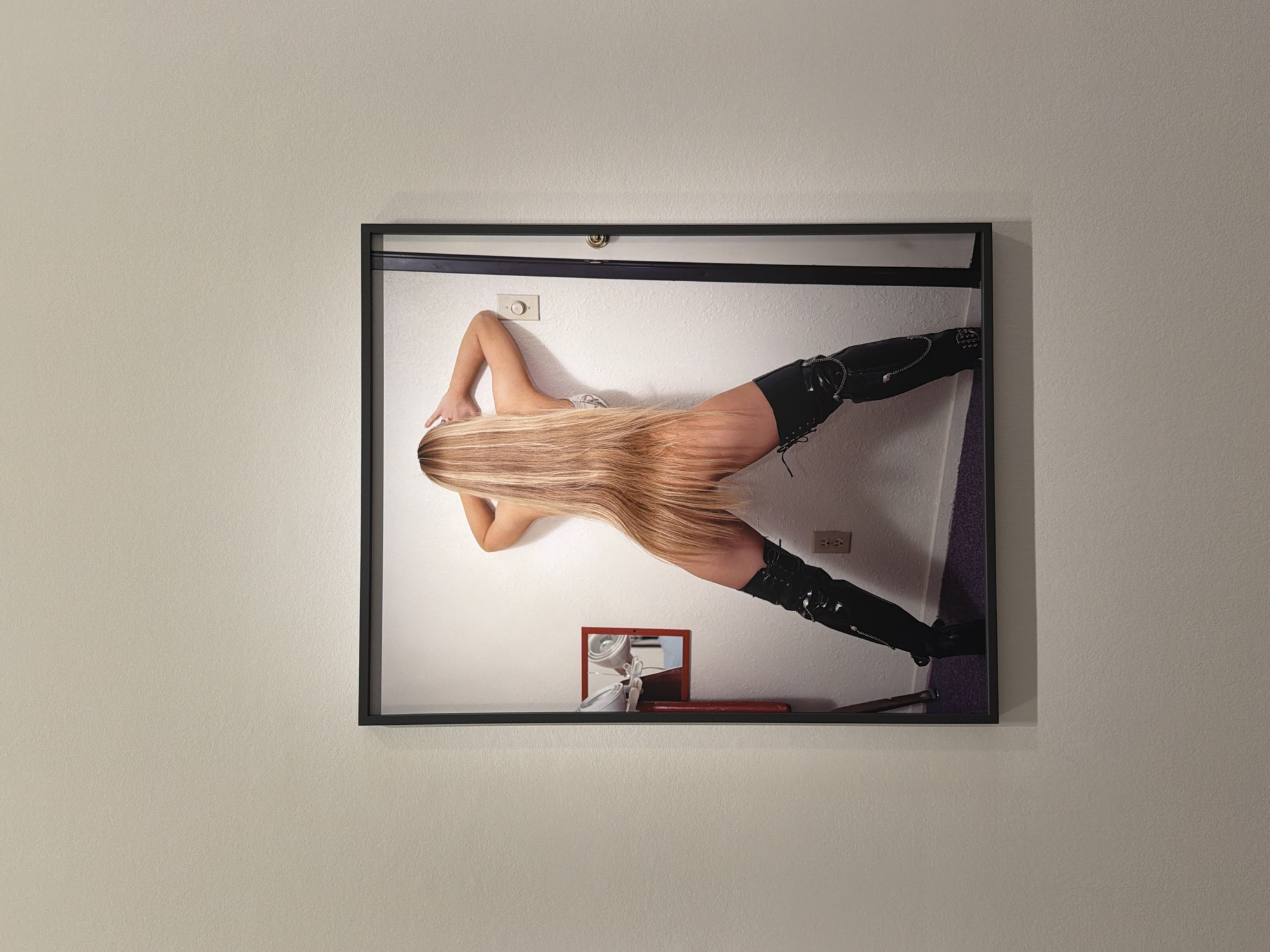 A large, black-framed artwork is prominently displayed on a plain, light-colored (likely off-white or cream) wall. The artwork itself features a person in a dynamic and unusual pose within an indoor setting.
The subject of the artwork, implied to be a female, has very long, straight blonde hair that cascades down her back, obscuring her face and upper torso. She is wearing black high-waisted shorts and striking black thigh-high or over-the-knee boots, which are laced up and adorned with chains and studs. A glimpse of a white top is visible. She is positioned against a white wall, appearing to lean or lie on it with her body angled downwards towards what looks like a purple carpet or rug. Her left arm is extended upwards, reaching towards a white light switch or dimmer on the wall. Below her, on a flat surface within the artwork's scene, a small mirror with a red frame reflects an indistinct white object, possibly a hair dryer, and a long, reddish item, perhaps a hairbrush. An electrical outlet is also visible on the white wall behind her lower leg.
The composition of the artwork creates a sense of defying gravity, as the person's body is oriented diagonally across the frame. There are no discernible emotions conveyed by the subject, and the time of day is indeterminate as the scene is indoors. No visible text is present in the image. This media file was taken in Paris, France.